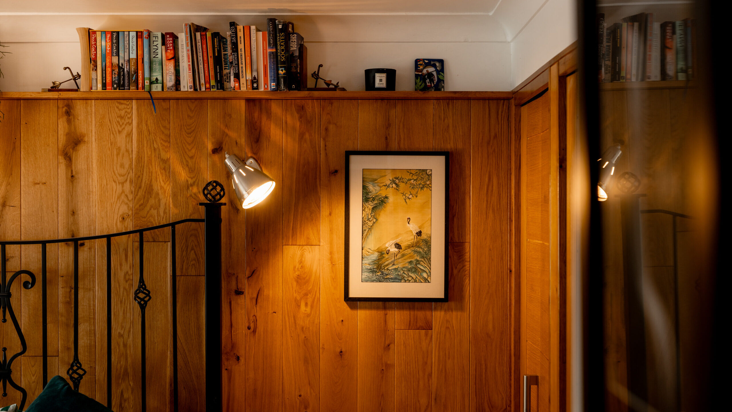 A cozy room with wooden walls, a framed painting, a metal bed frame, a wall-mounted lamp illuminating the artwork, and a bookshelf filled with books and small decor items above.
