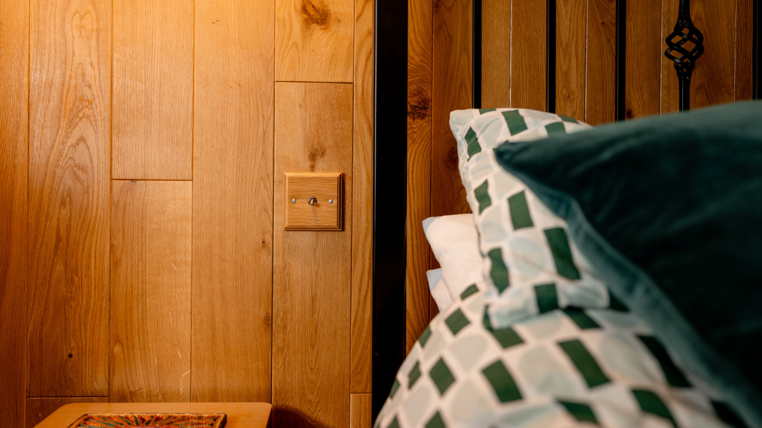 A close-up of a cozy bed with geometric-patterned bedding, next to a wooden wall with a light switch and a small bedside table holding a colorful coaster. Warm lighting creates a relaxing atmosphere.
