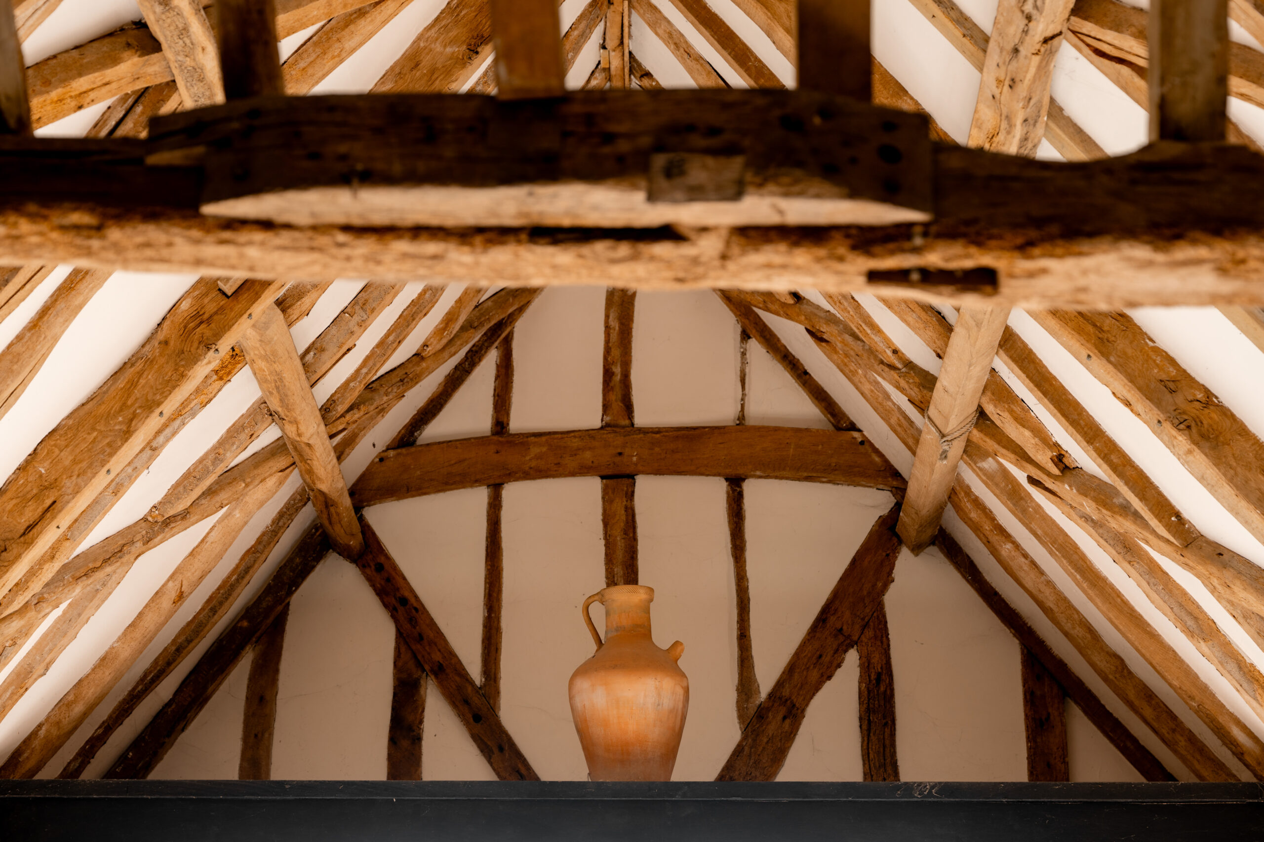 A clay jug is placed on a black shelf beneath a rustic wooden beam ceiling, featuring exposed wooden beams and white plaster in a loft or attic space.