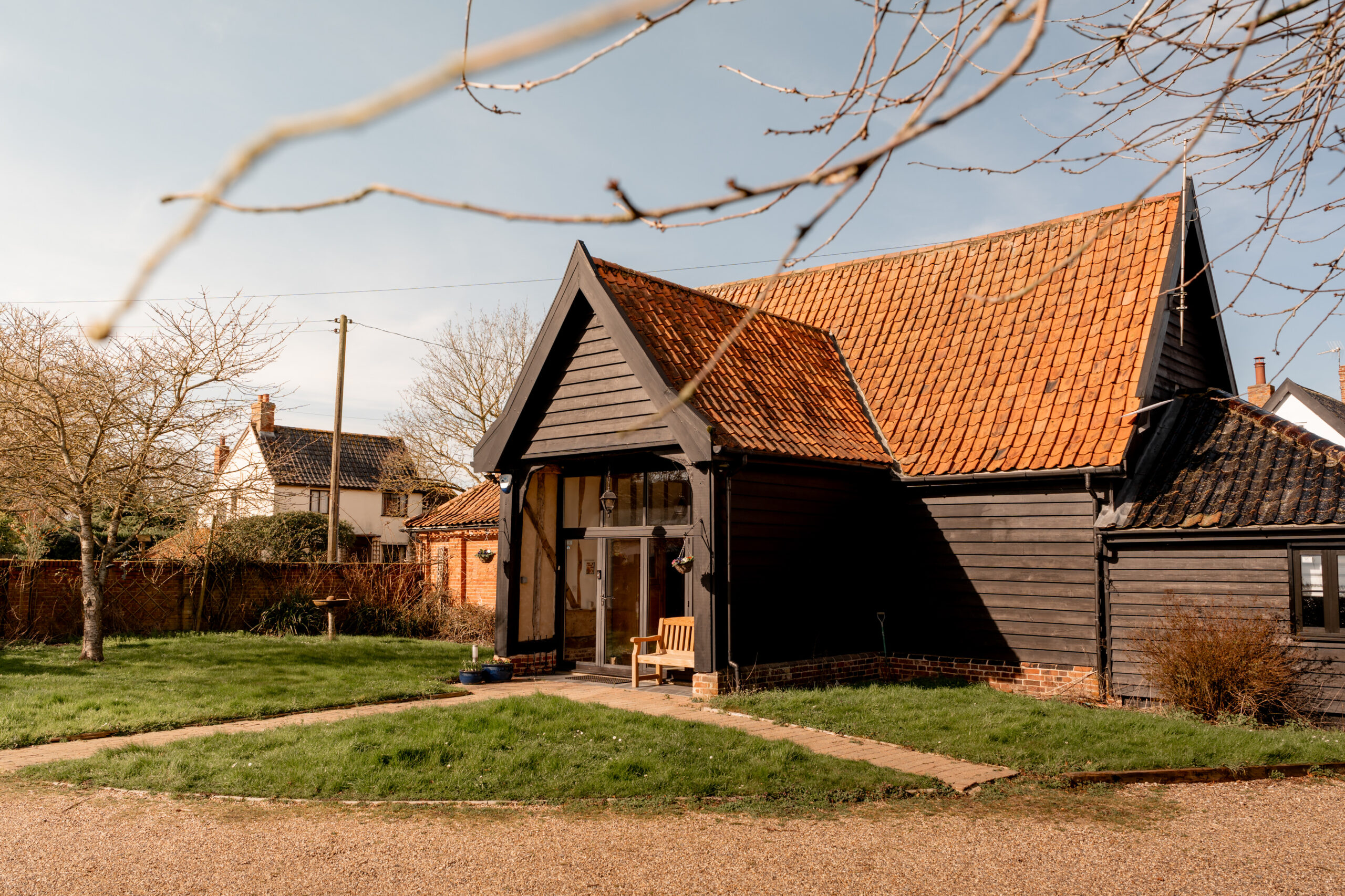 A modern barn-style house with dark wood siding and a red tiled roof sits on a grassy yard. A wooden bench is by the glass entrance, and there are leafless trees and other houses in the background.