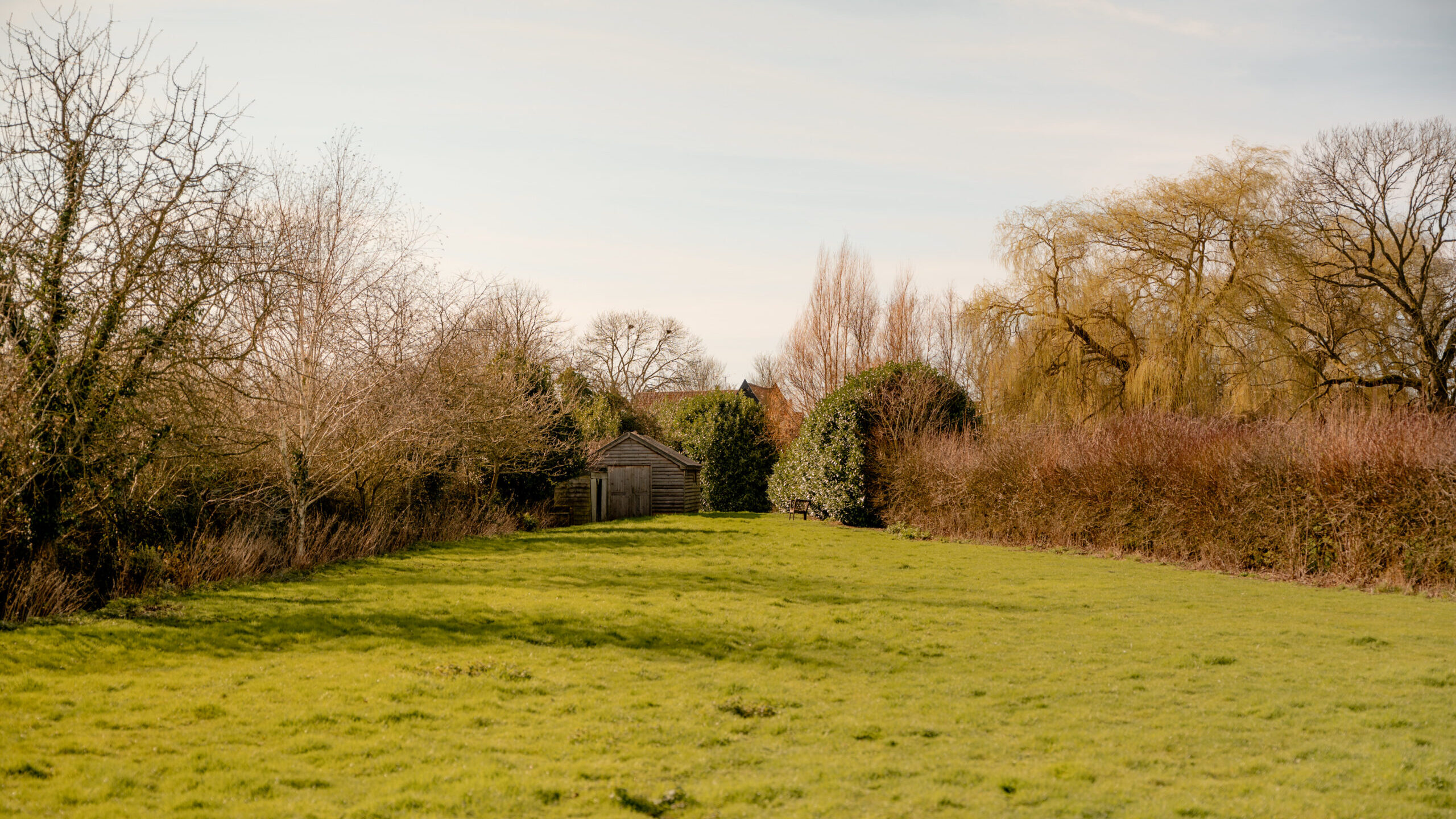 A grassy field bordered by leafless trees and bushes, with a wooden shed partially hidden among tall trees in the background under a partly cloudy sky.