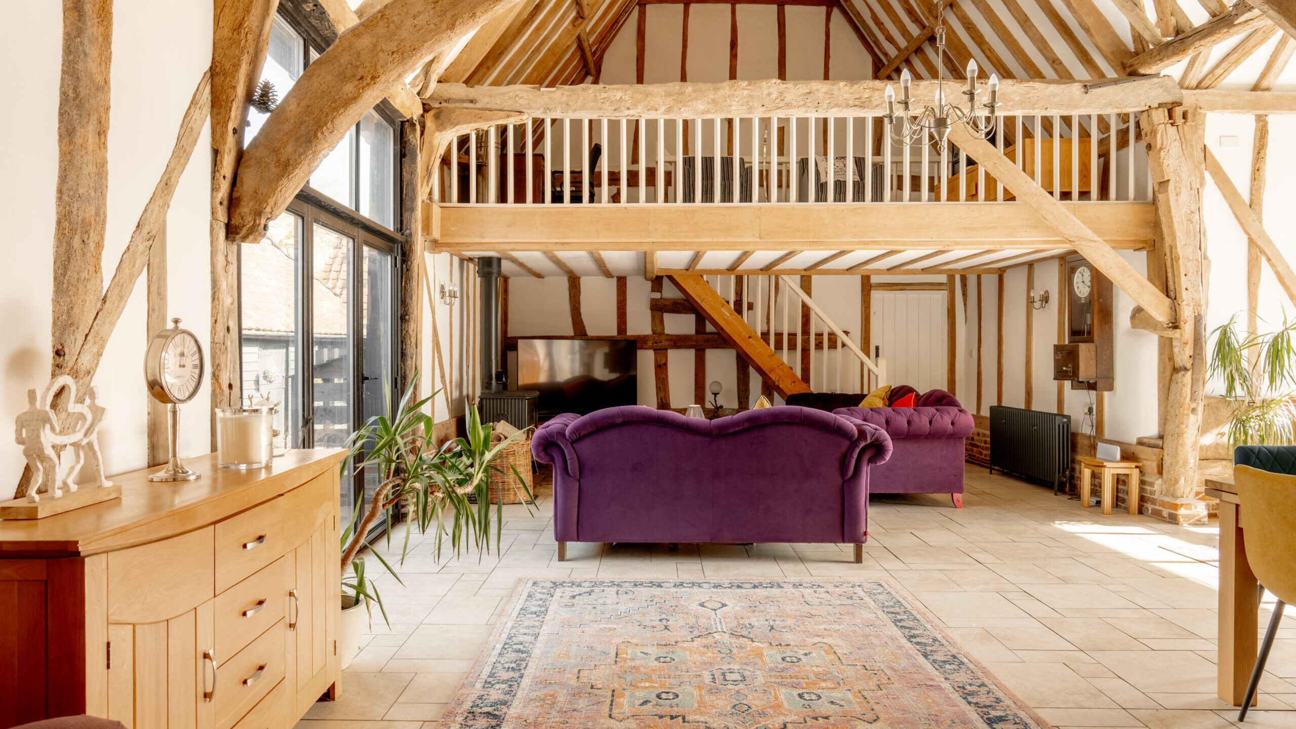 A spacious living room with exposed wooden beams, a vaulted ceiling, and a mezzanine balcony. There are purple sofas, a TV, large windows, a chandelier, and a patterned rug on a tiled floor.