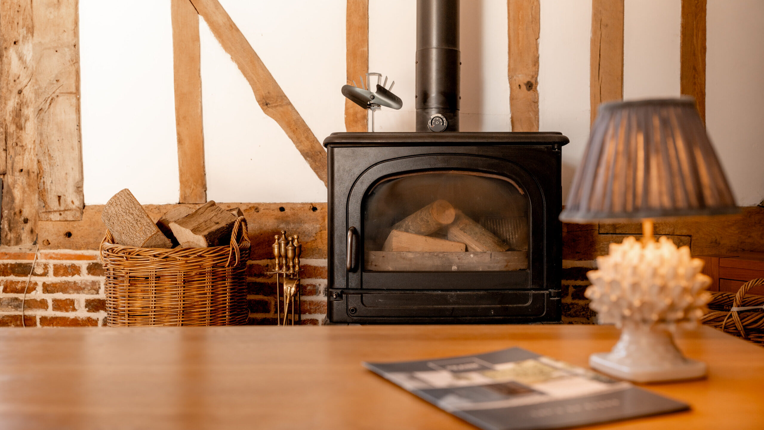 A cozy room with a black wood-burning stove, stacked firewood in a wicker basket, and a lit table lamp on a wooden surface, with magazines on the table and a rustic timber-framed wall in the background.