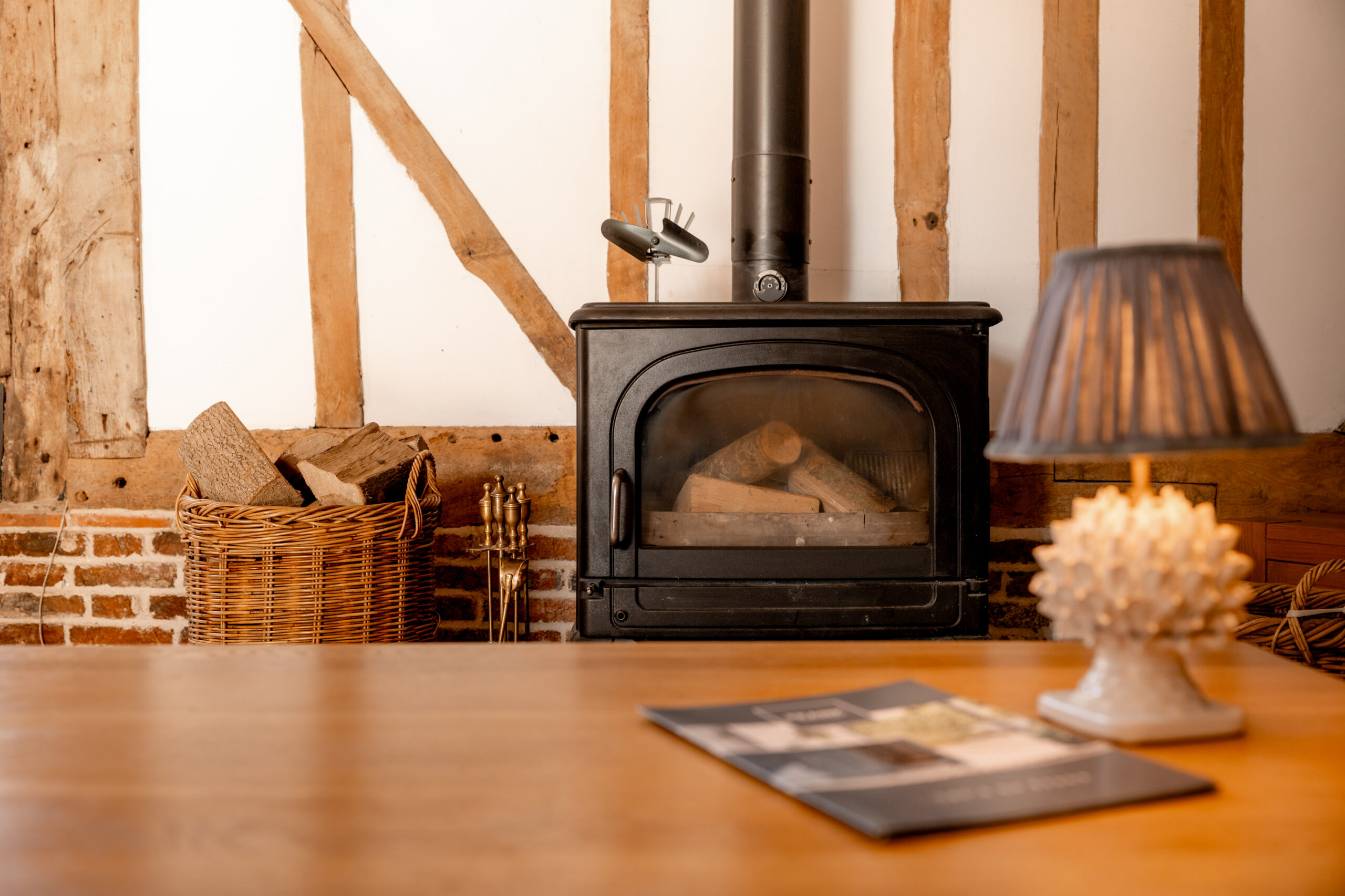 A cozy room with a black wood-burning stove, stacked firewood in a wicker basket, and a lit table lamp on a wooden surface, with magazines on the table and a rustic timber-framed wall in the background.
