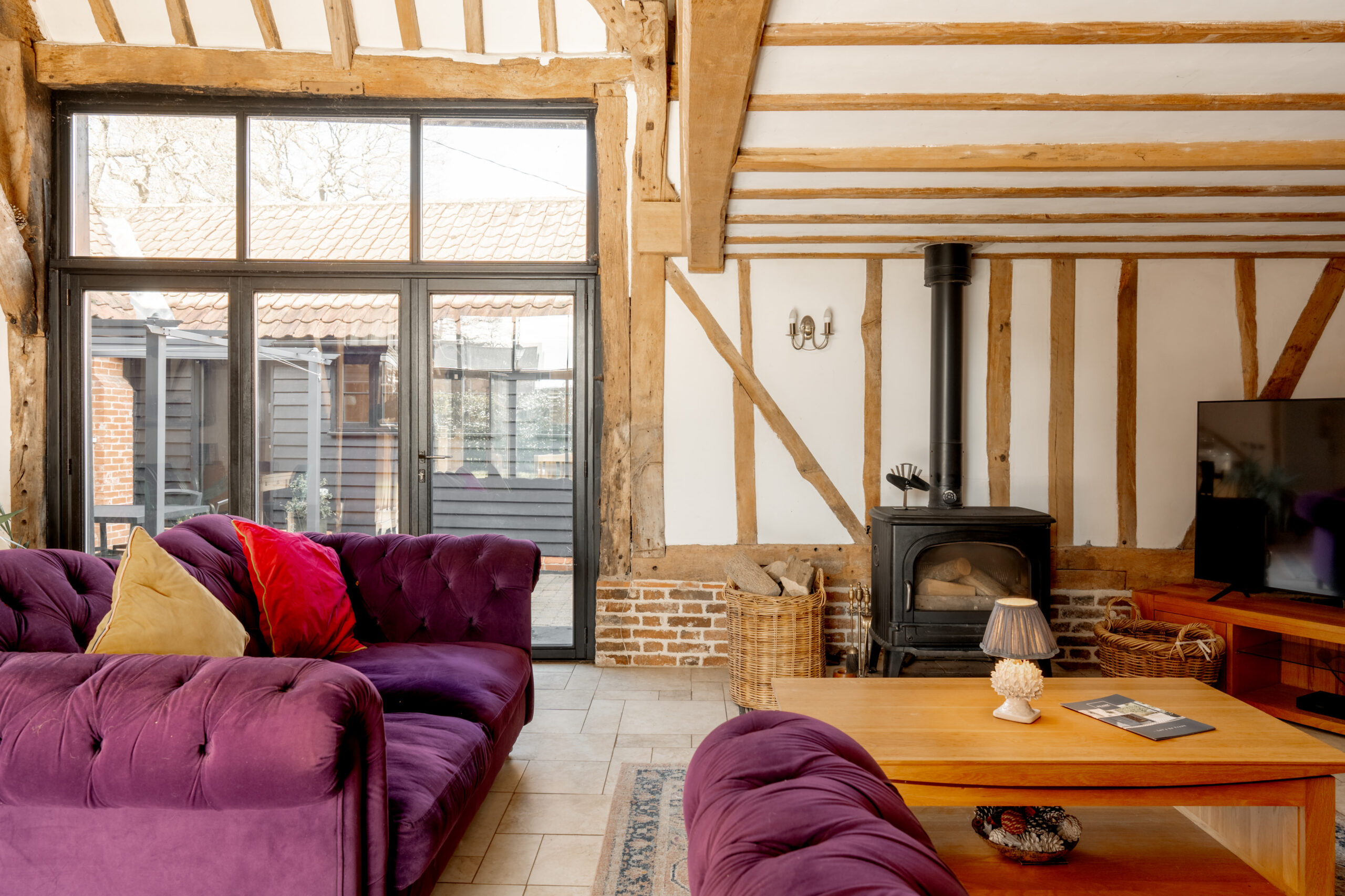 A cozy living room with exposed wooden beams, large windows, purple tufted sofas, a wood-burning stove, a wooden coffee table, and a TV on a wooden stand. Natural light fills the rustic space.
