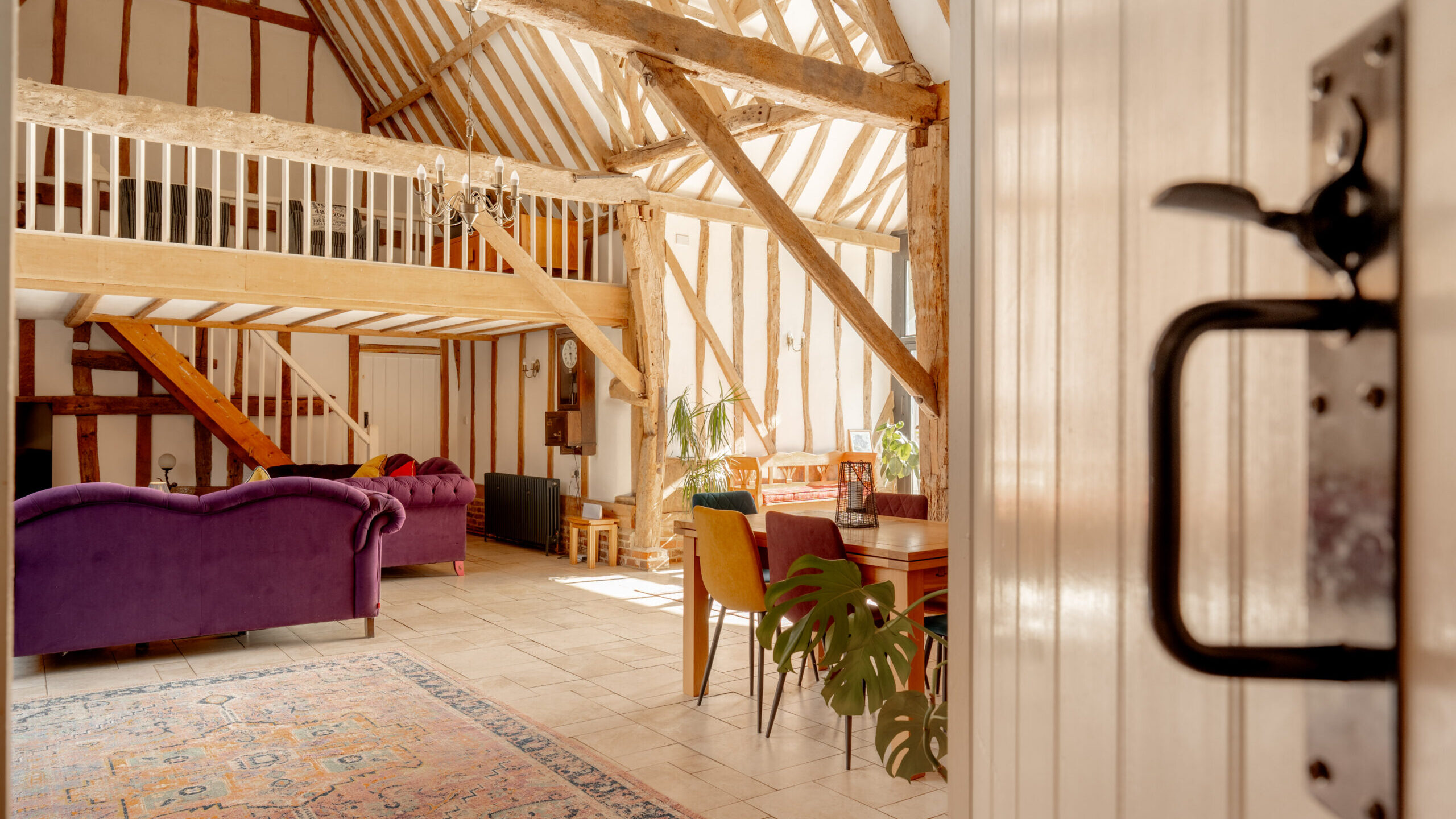 Sunlit, rustic living space with exposed wooden beams, loft area, purple sofas, dining table with plants, colorful rug, and tiled floor. Photo is taken from an open door with a black handle in the foreground.