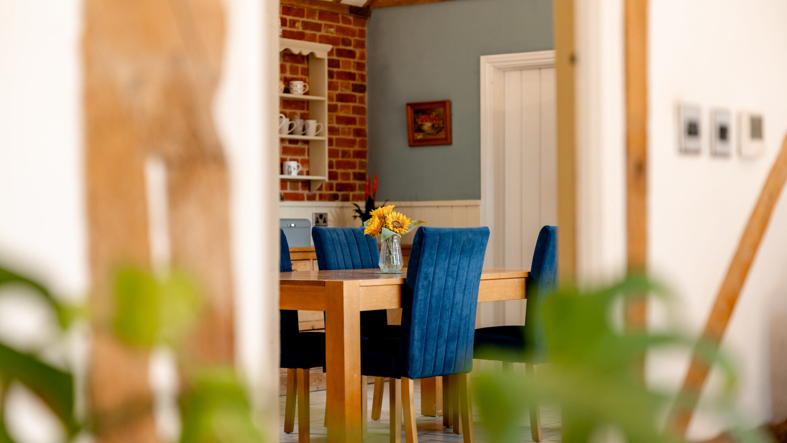 A wooden dining table with four blue upholstered chairs and a vase of sunflowers sits in a cozy, sunlit room with exposed beams, brick accents, and a blue painted wall in the background.