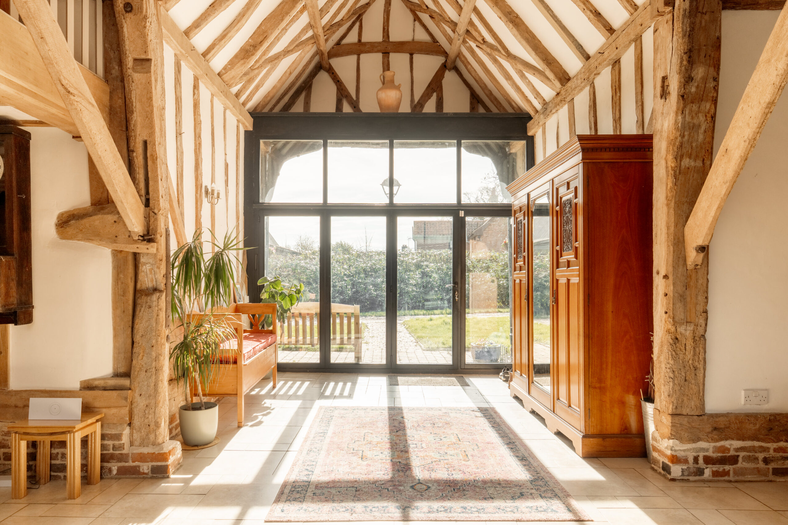 A bright, rustic hallway with exposed wooden beams, large glass doors leading to a garden, a wooden bench with plants, an ornate rug, and a vintage wooden wardrobe. Sunlight fills the space.