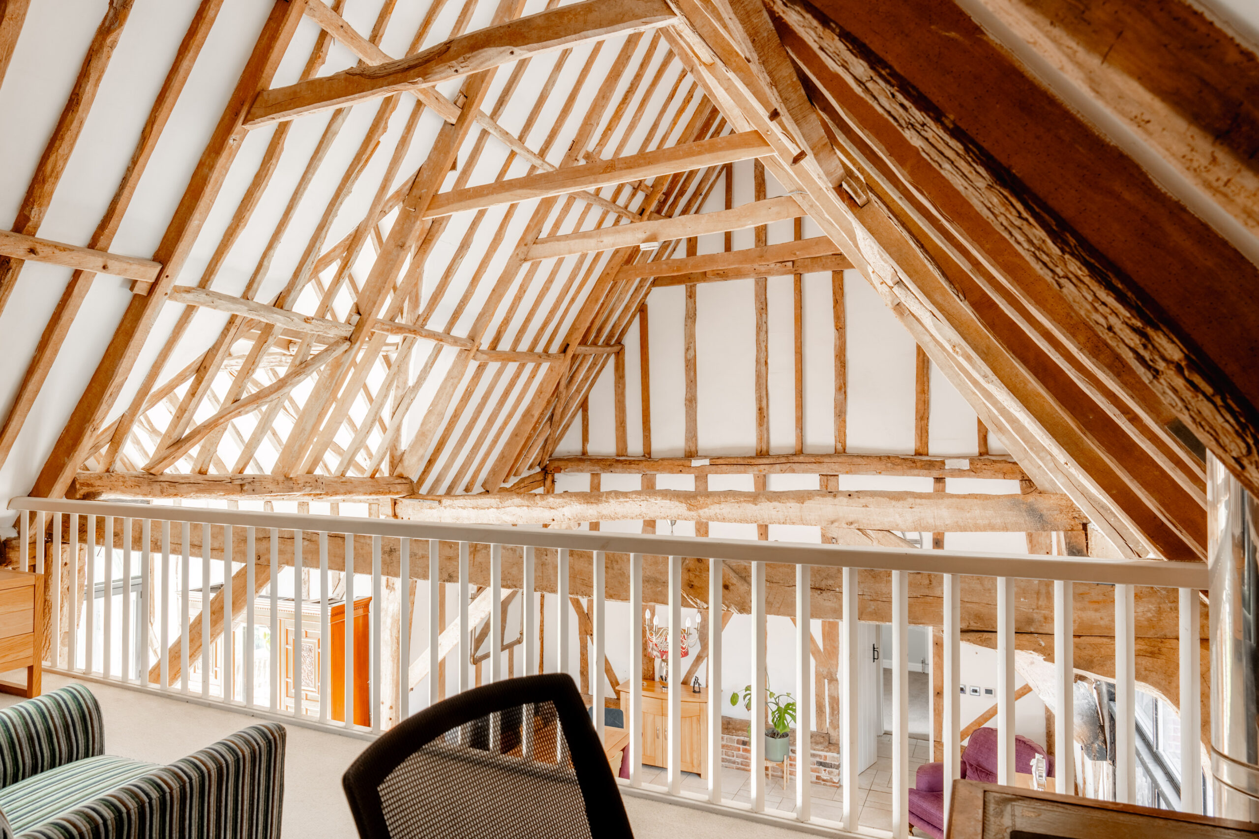 A loft space with exposed wooden beams and white walls, overlooking a lower level. The area features a railing, striped sofa, and modern chair, highlighting the rustic and airy architectural design.