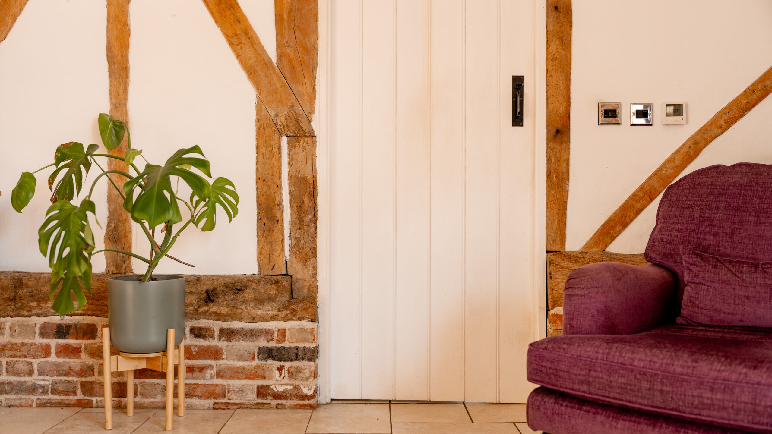 A potted monstera plant on a wooden stand sits beside a white paneled door, with exposed wooden beams and brickwork on the wall. A purple armchair is partially visible to the right.