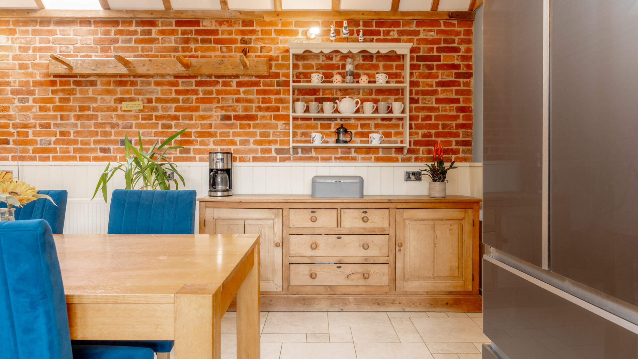 A cozy kitchen with exposed brick walls, wooden cabinets, blue upholstered chairs around a wooden table, a coffee maker, plants, and a white shelf holding cups and teapots.