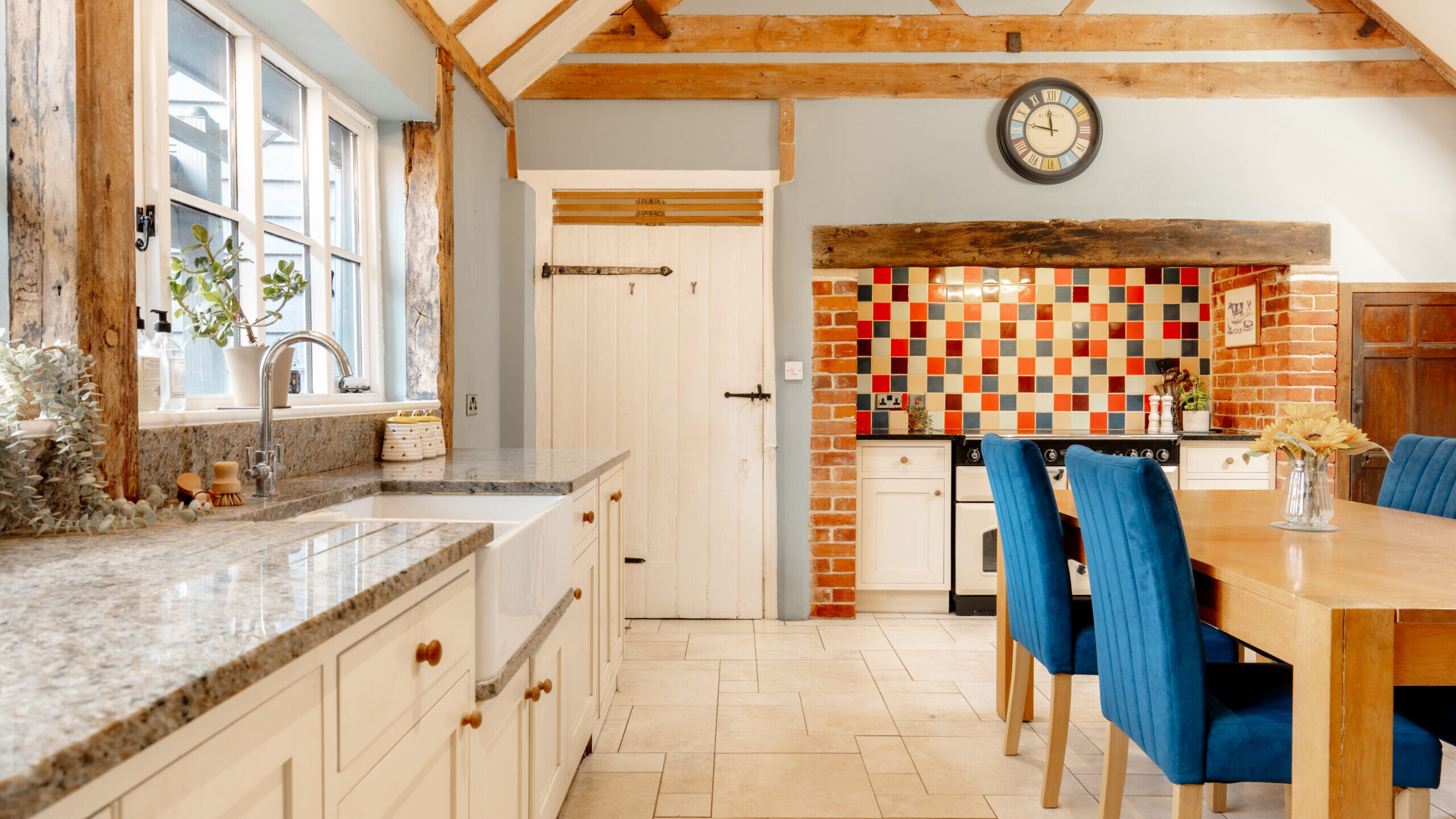 A bright rustic kitchen features exposed wooden beams, a farmhouse sink, and a granite countertop. Blue chairs surround a wooden dining table, and a colorful tiled backsplash adds vibrancy near the stove area.