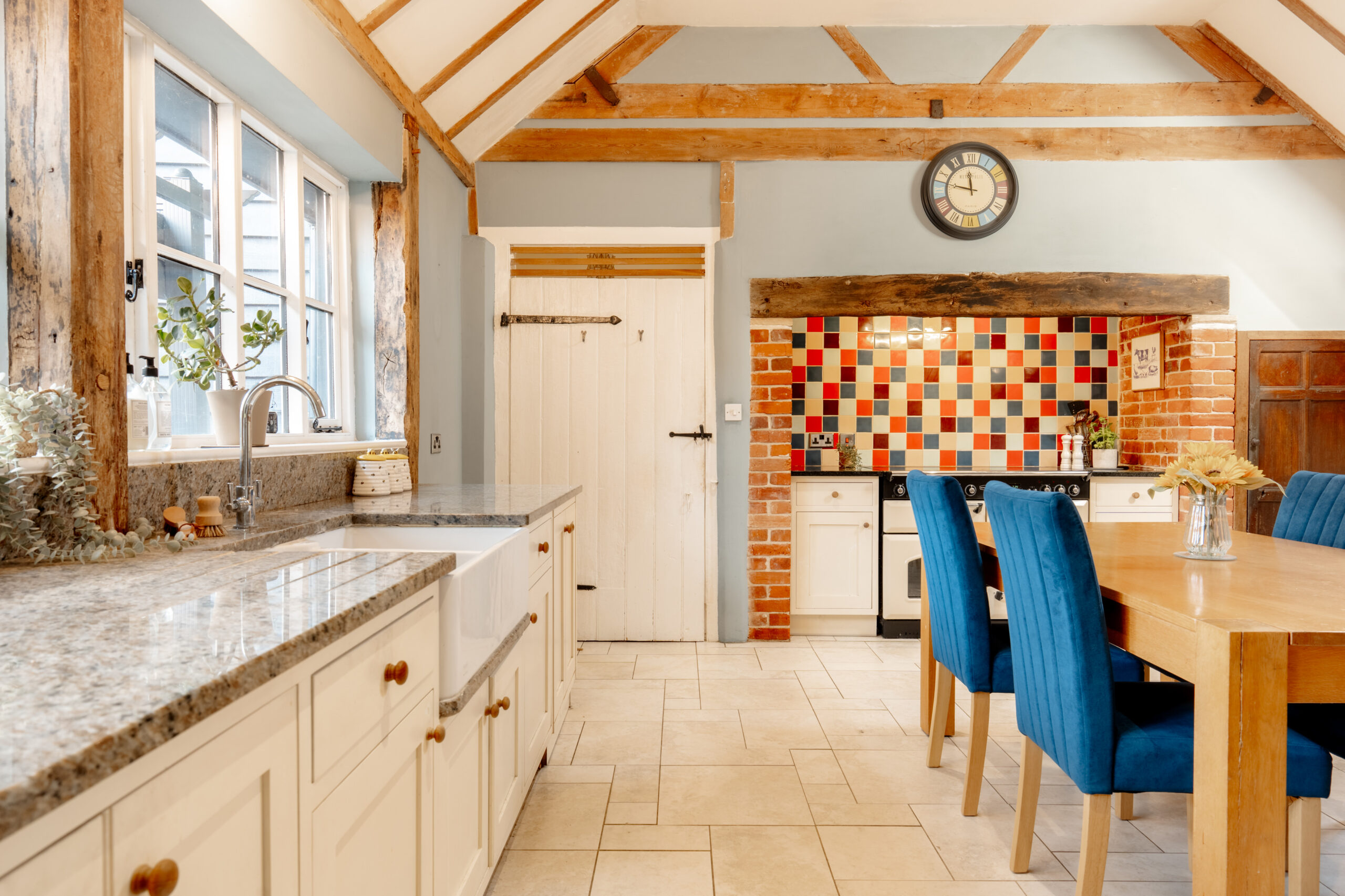 A bright rustic kitchen features exposed wooden beams, a farmhouse sink, and a granite countertop. Blue chairs surround a wooden dining table, and a colorful tiled backsplash adds vibrancy near the stove area.