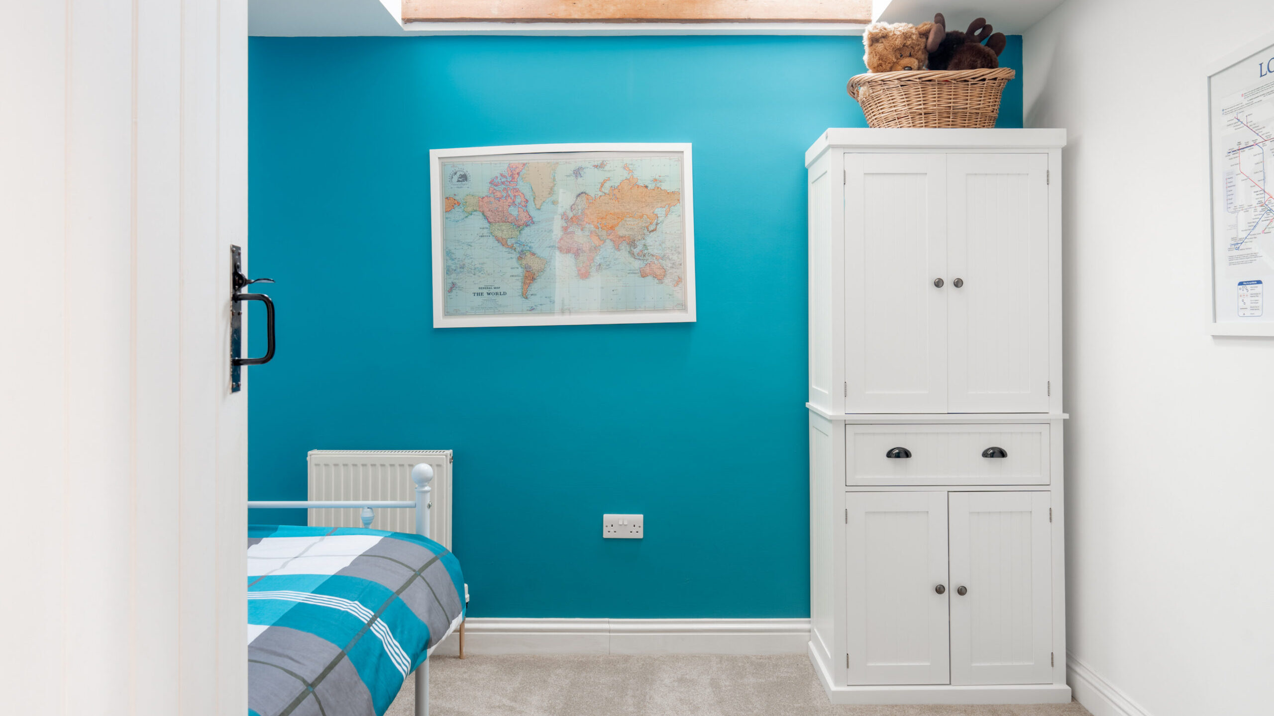 A bright bedroom with a skylight, teal accent wall, world map, white wardrobe, and a single bed with blue plaid bedding. A basket with stuffed animals sits on top of the wardrobe.
