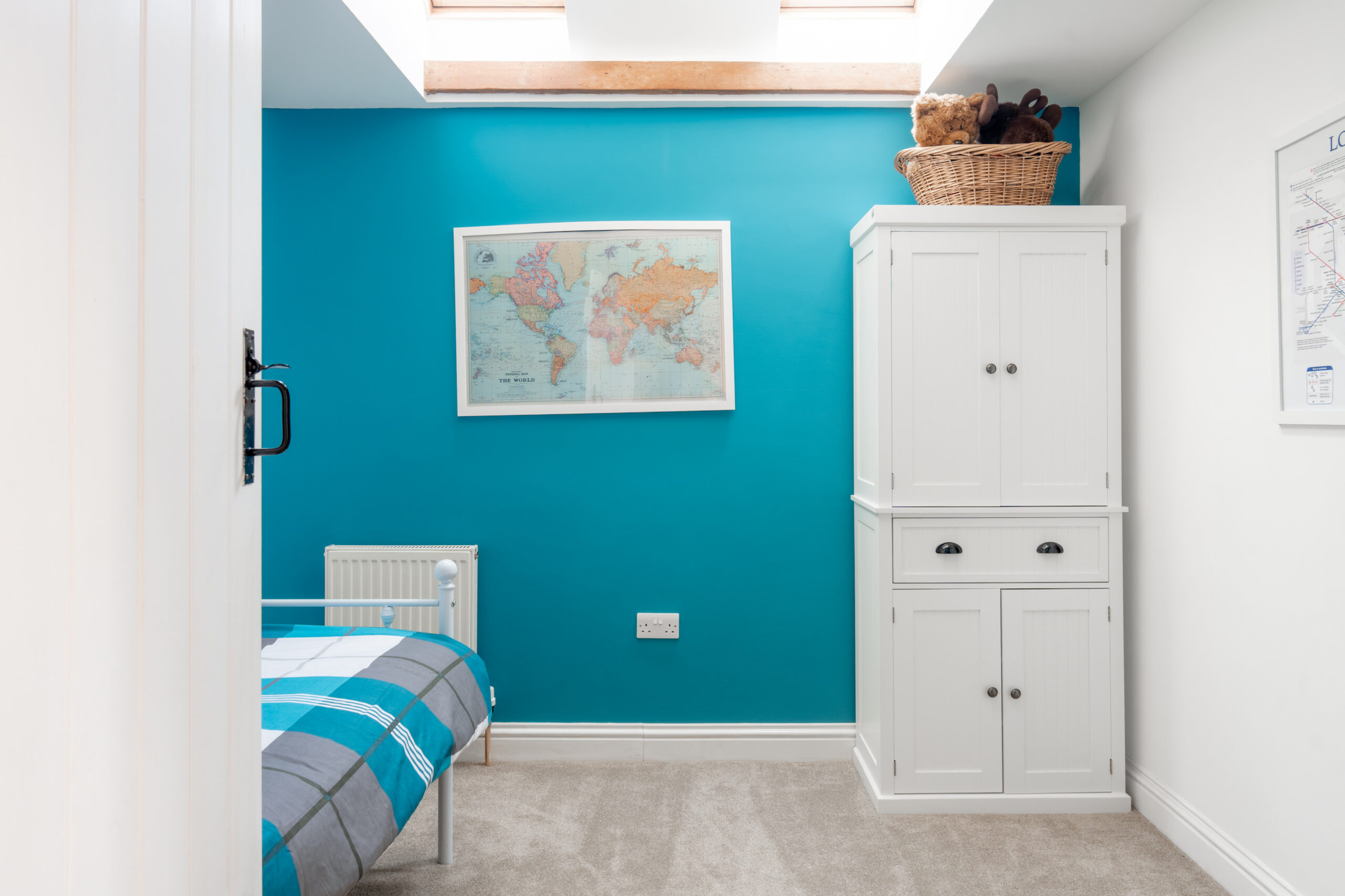 A bright bedroom with a skylight, teal accent wall, world map, white wardrobe, and a single bed with blue plaid bedding. A basket with stuffed animals sits on top of the wardrobe.