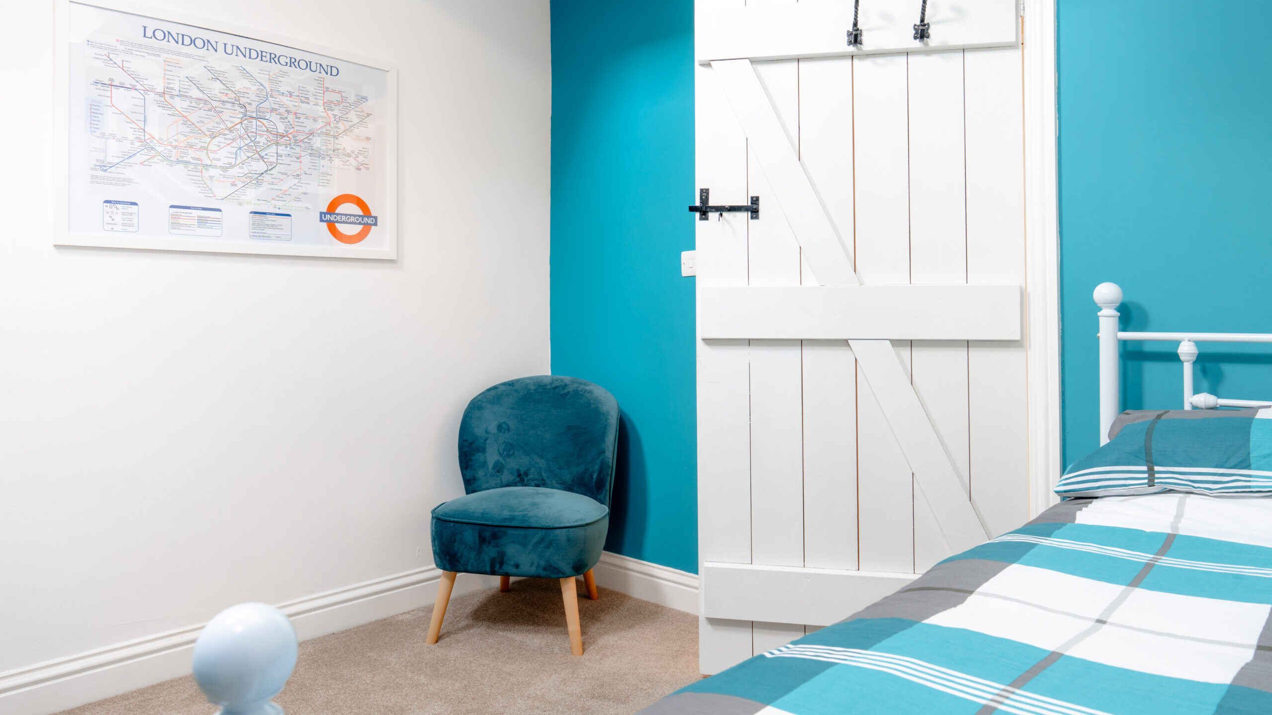 A bright bedroom with turquoise and white walls, a white bed with blue and gray checkered bedding, a teal velvet chair, a barn-style door, and a framed London Underground map on the wall.