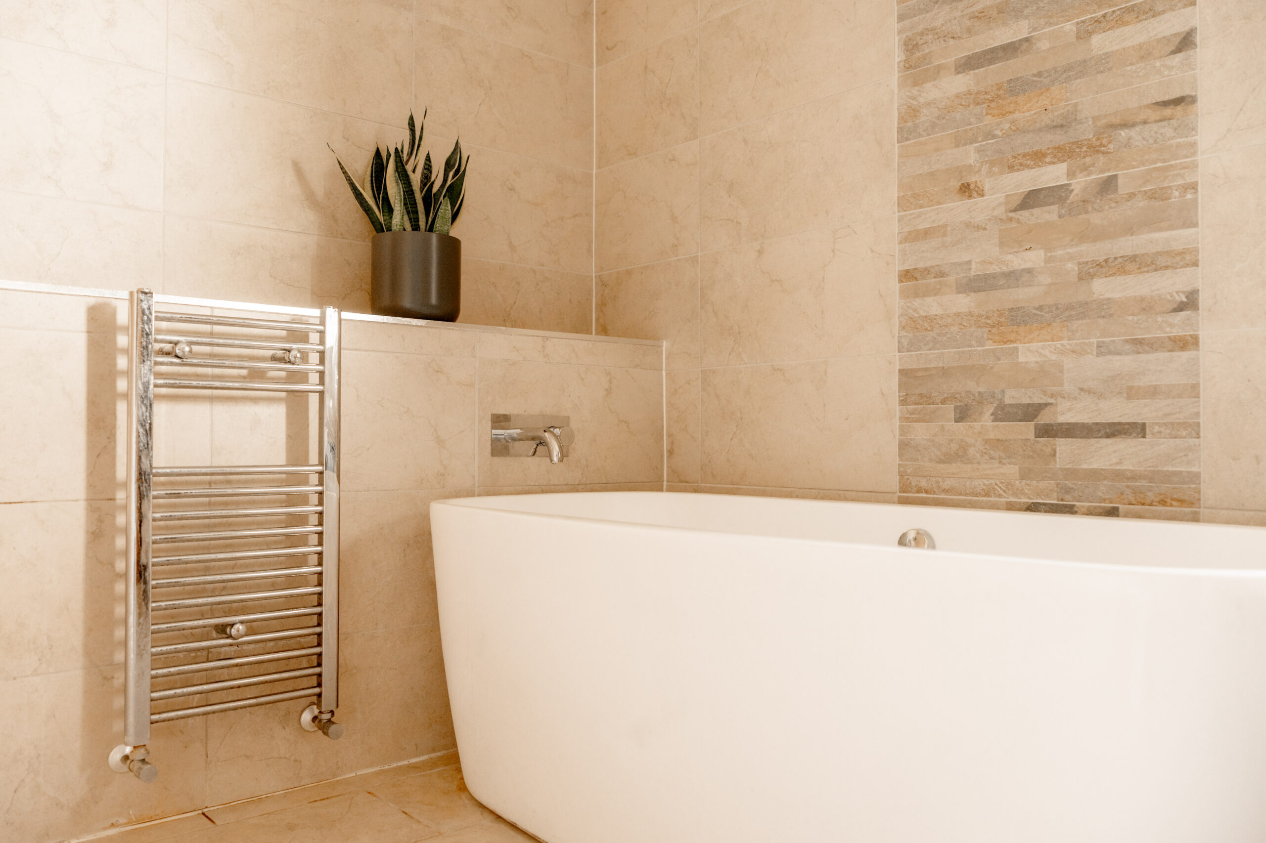 Modern bathroom with beige tiled walls, a white bathtub, a chrome towel warmer, and a potted snake plant on a built-in shelf. The wall behind the tub features a decorative stone tile accent.