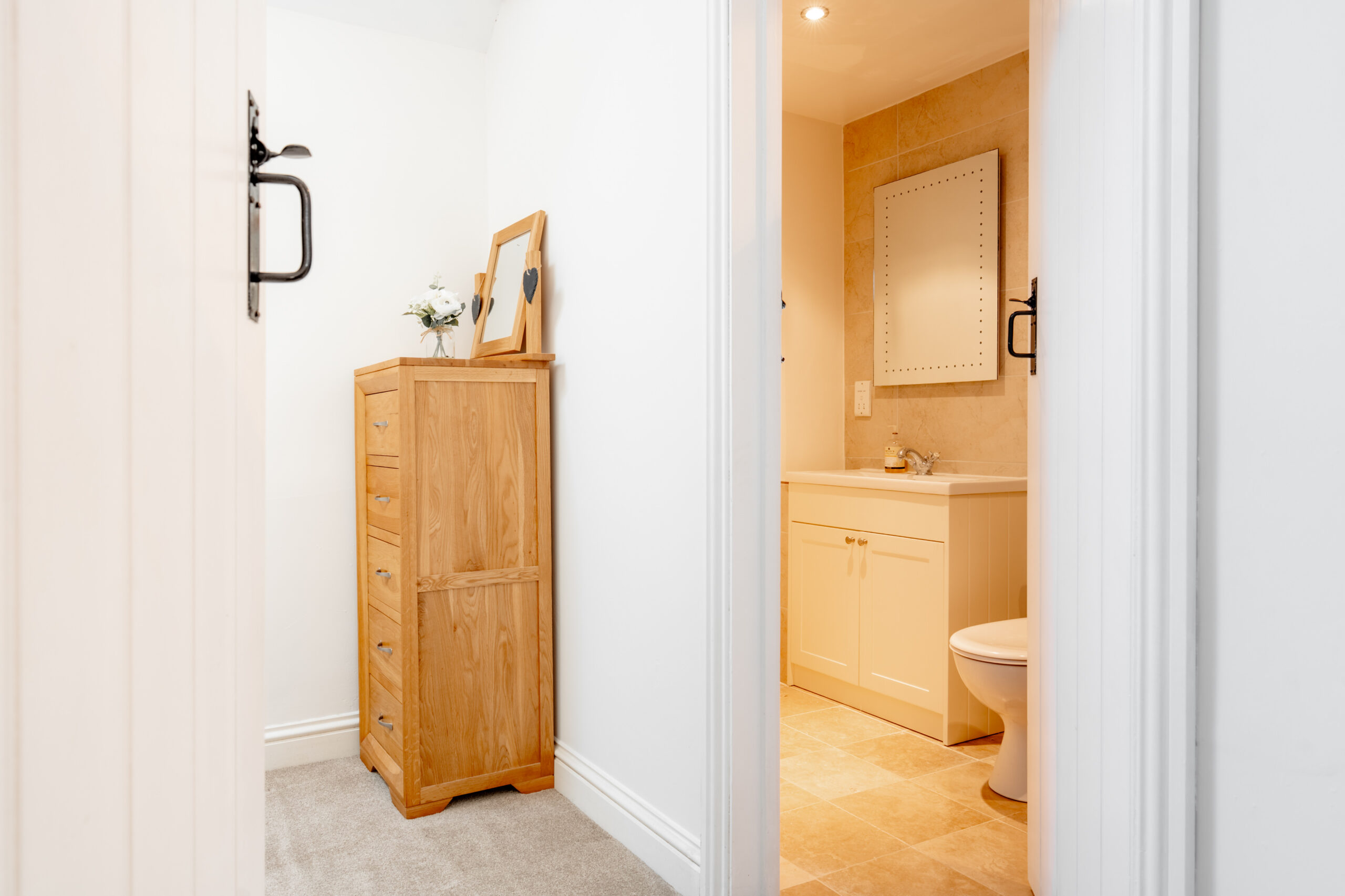 A view through an open doorway reveals a light, modern bathroom with a white vanity and tan tiles. Outside the bathroom is a small area with a wooden dresser, framed photo, and a small vase with flowers.