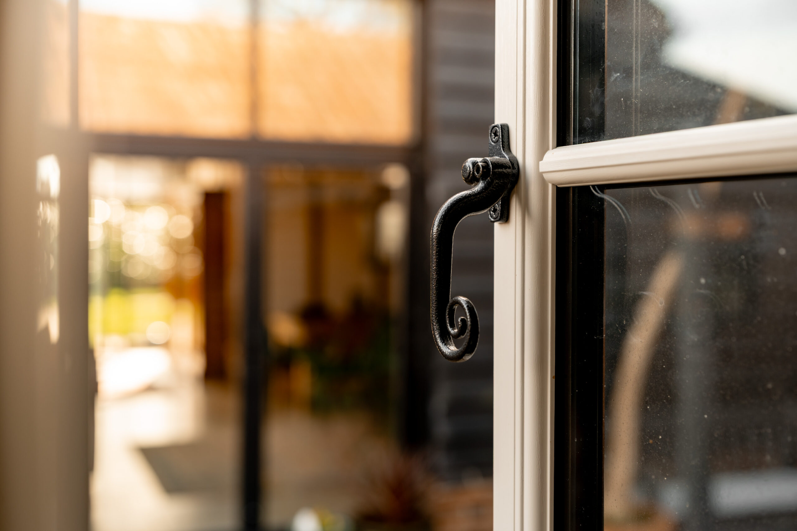 Close-up of a black, ornate handle on an open window, with a blurred view of a sunlit indoor space and garden beyond in the background.