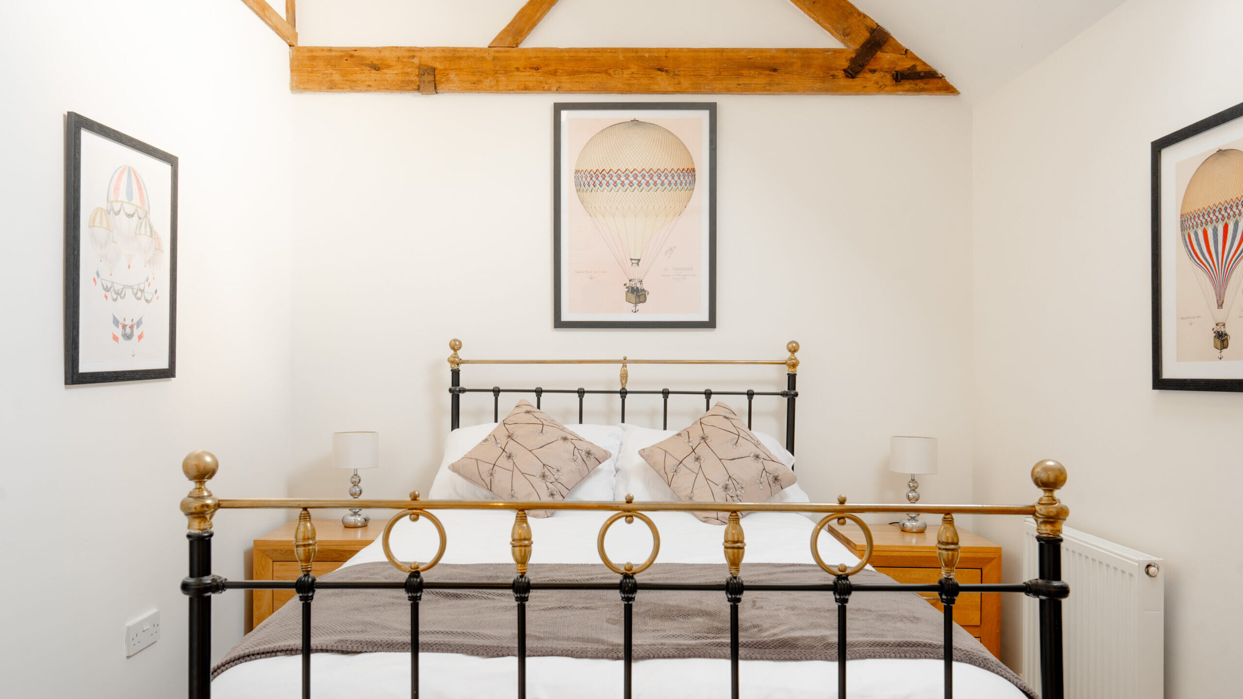 A cozy bedroom with a black metal bed frame, white bedding, and two map-print pillows. Hot air balloon artwork decorates the white walls, and wooden beams are visible on the ceiling. Two bedside tables with lamps flank the bed.