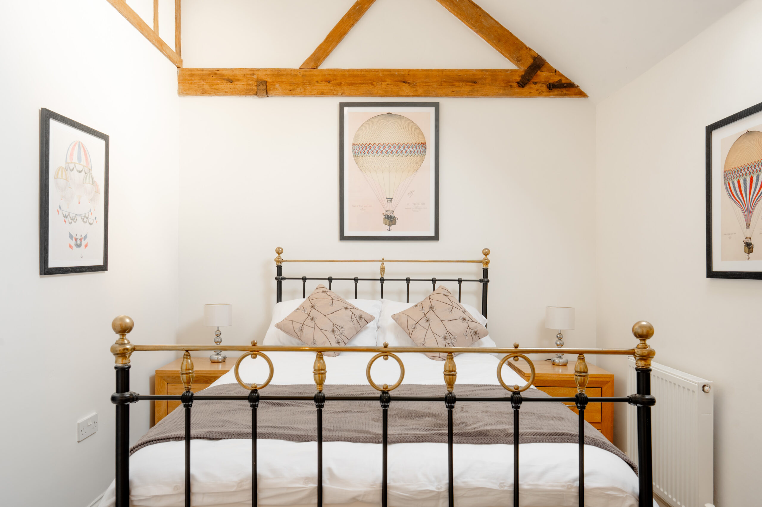 A cozy bedroom with a black metal bed frame, white bedding, and two map-print pillows. Hot air balloon artwork decorates the white walls, and wooden beams are visible on the ceiling. Two bedside tables with lamps flank the bed.