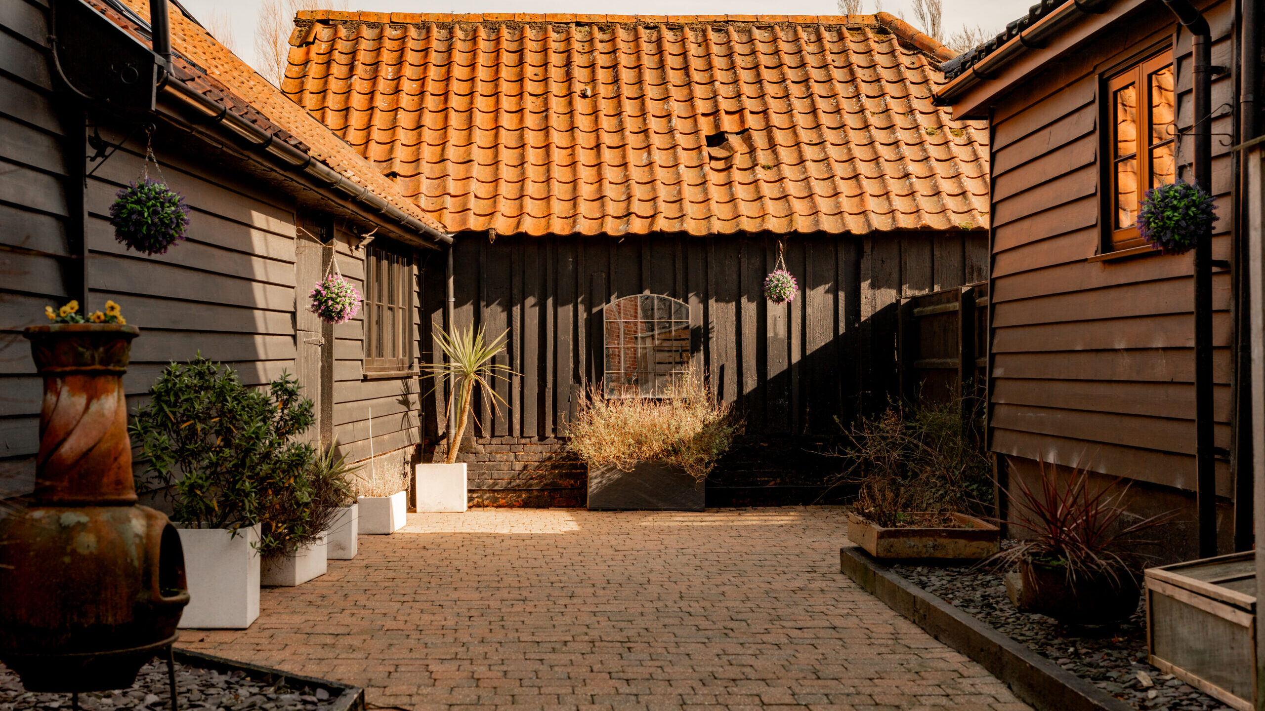 A brick pathway leads between two dark wooden buildings toward a shed with a red tile roof. Potted plants and hanging baskets line the path, and sunlight casts warm shadows across the courtyard.