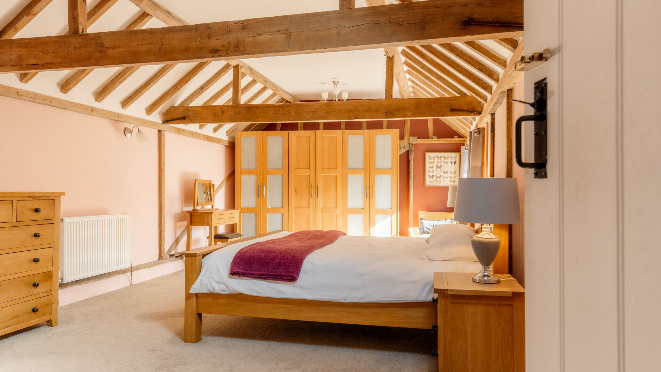 Spacious bedroom with wooden beams, a double bed with a white duvet and purple throw, wooden furniture, a bedside lamp, and large wardrobes along the back wall. Soft lighting and pastel pink walls create a cozy atmosphere.