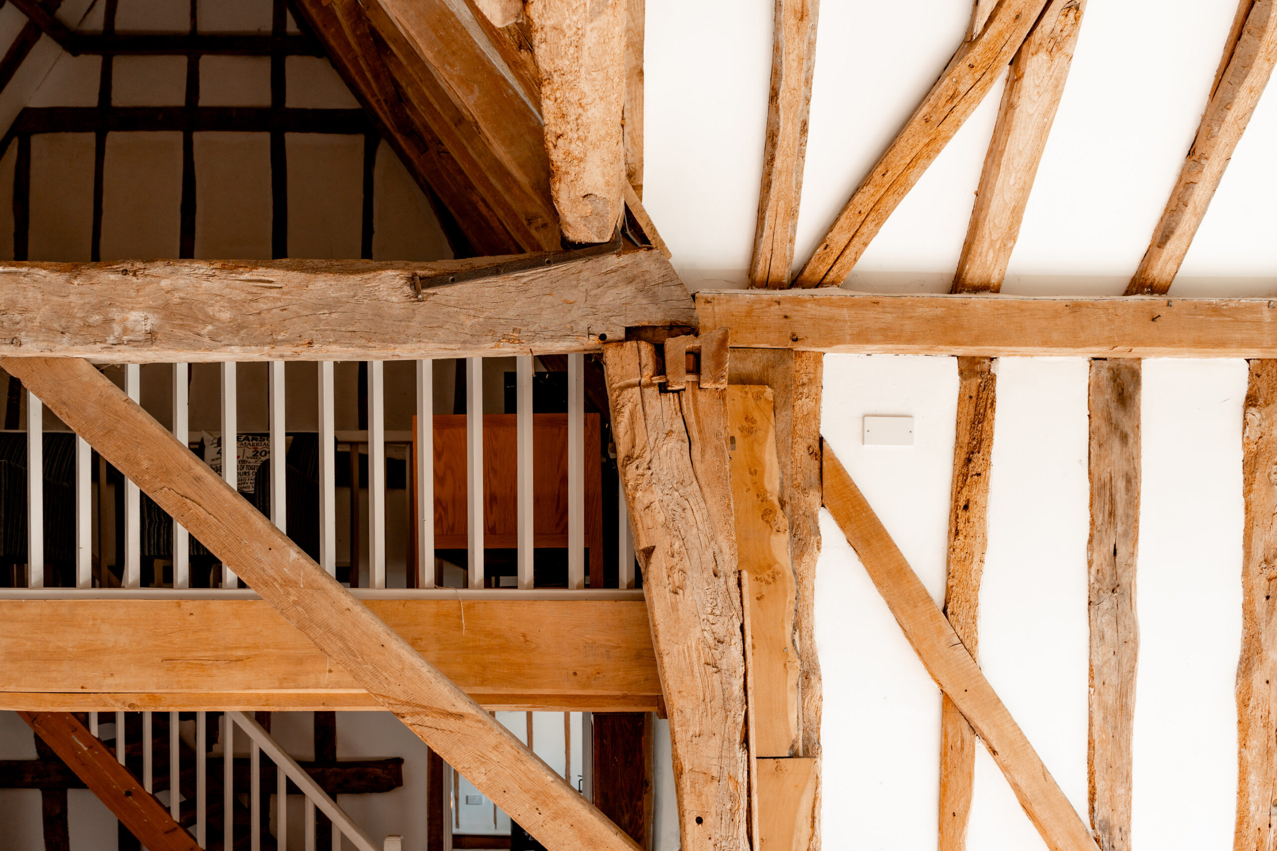 Exposed wooden beams intersect on a white wall in a rustic interior, with a railing and part of a wooden staircase visible in the background. The structure highlights traditional timber framing.