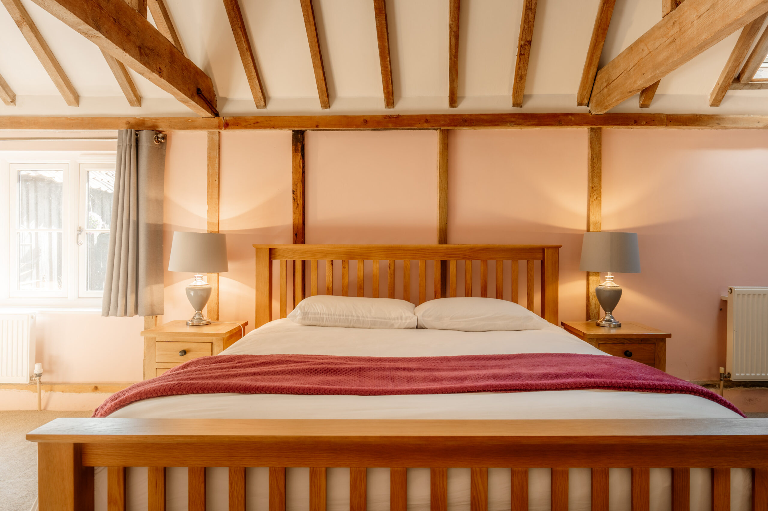 A cozy bedroom with a wooden bed frame, white bedding, and a pink throw blanket. Two bedside tables with matching lamps sit on either side, and wooden ceiling beams add rustic charm to the room. Light streams in from a window.