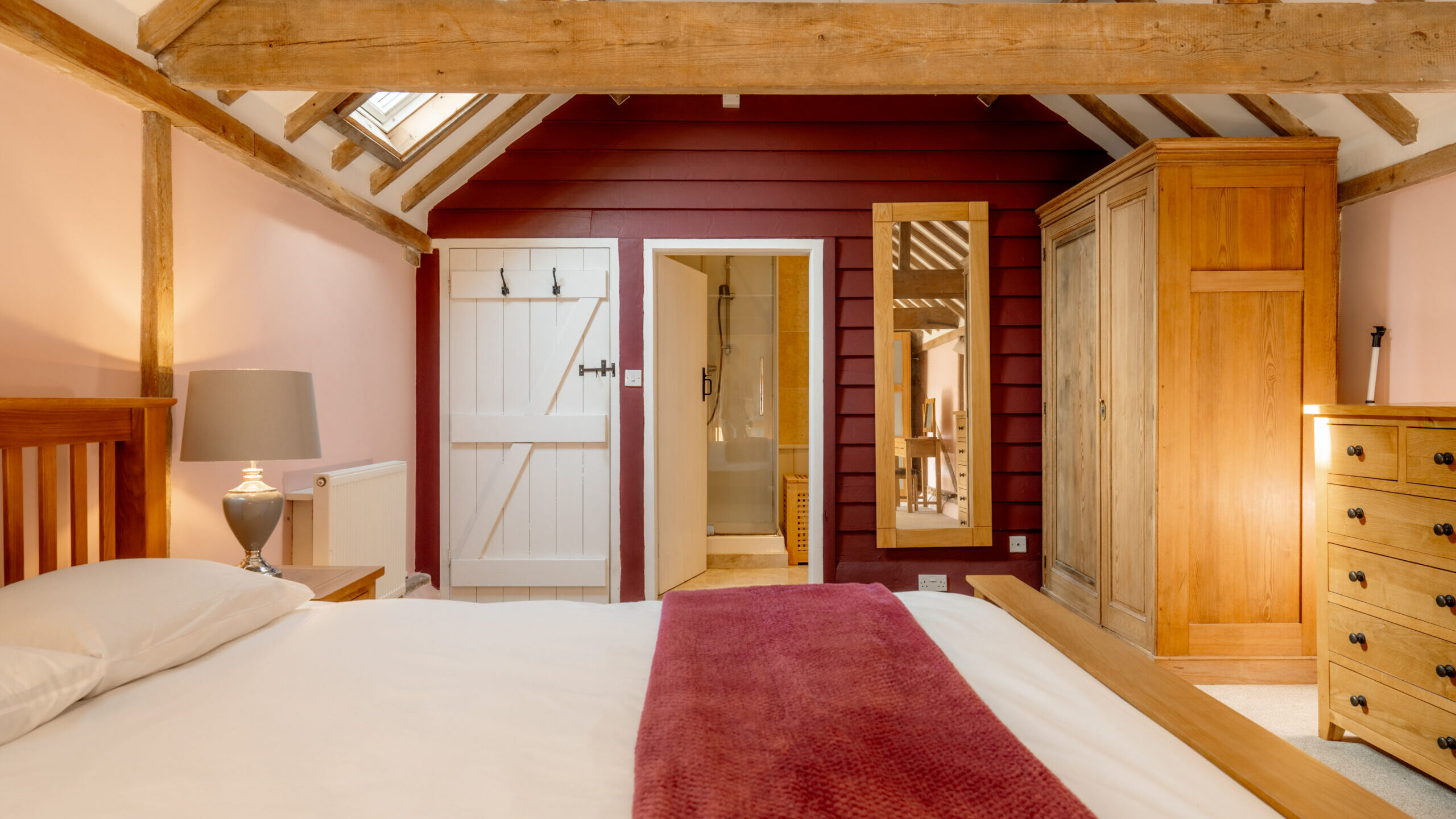Cozy bedroom with exposed wooden beams, a bed with white linens and a maroon throw, wooden furniture, a mirror, a lamp, and an open door leading to a bright bathroom. Warm lighting creates an inviting atmosphere.