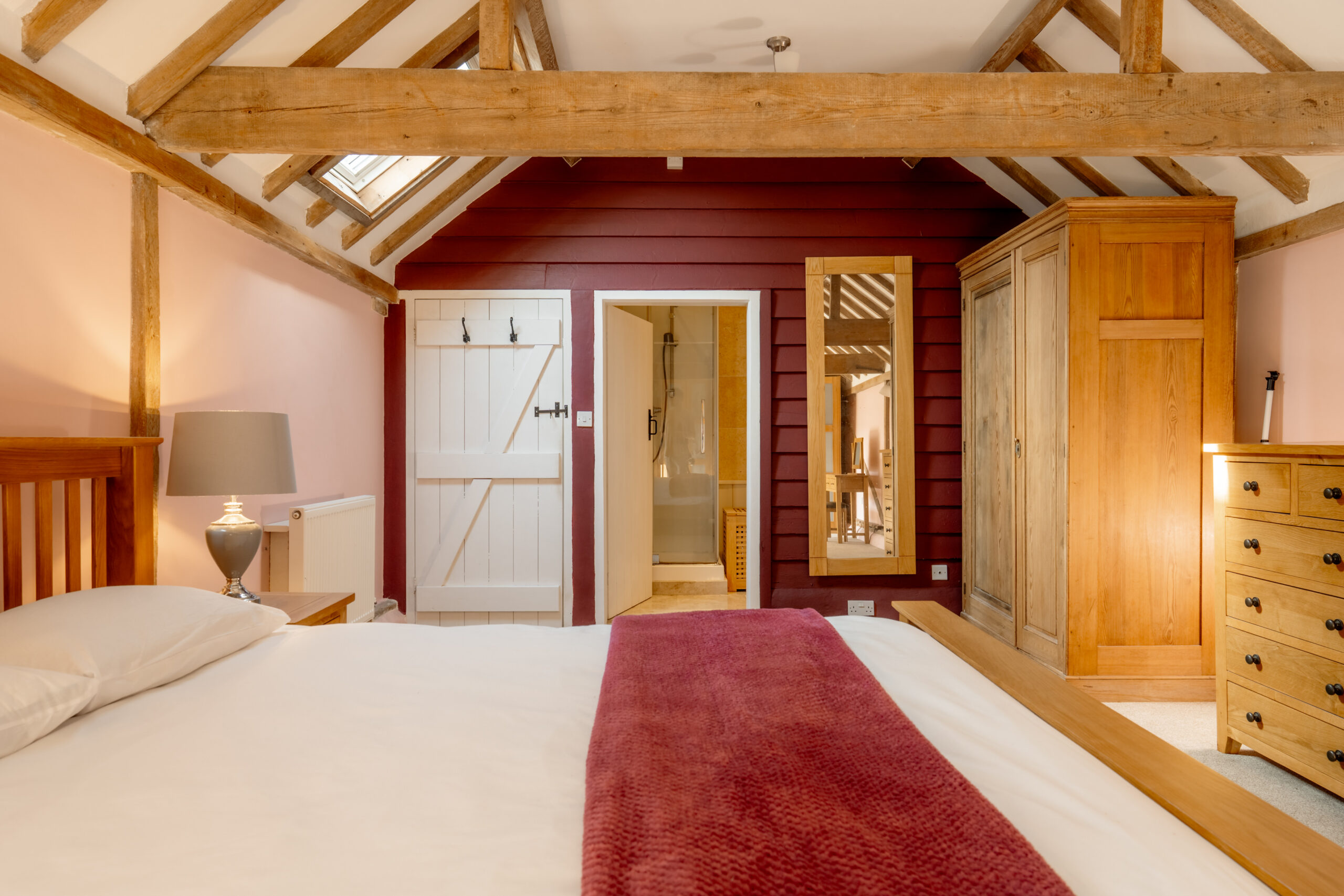 Cozy bedroom with exposed wooden beams, a bed with white linens and a maroon throw, wooden furniture, a mirror, a lamp, and an open door leading to a bright bathroom. Warm lighting creates an inviting atmosphere.