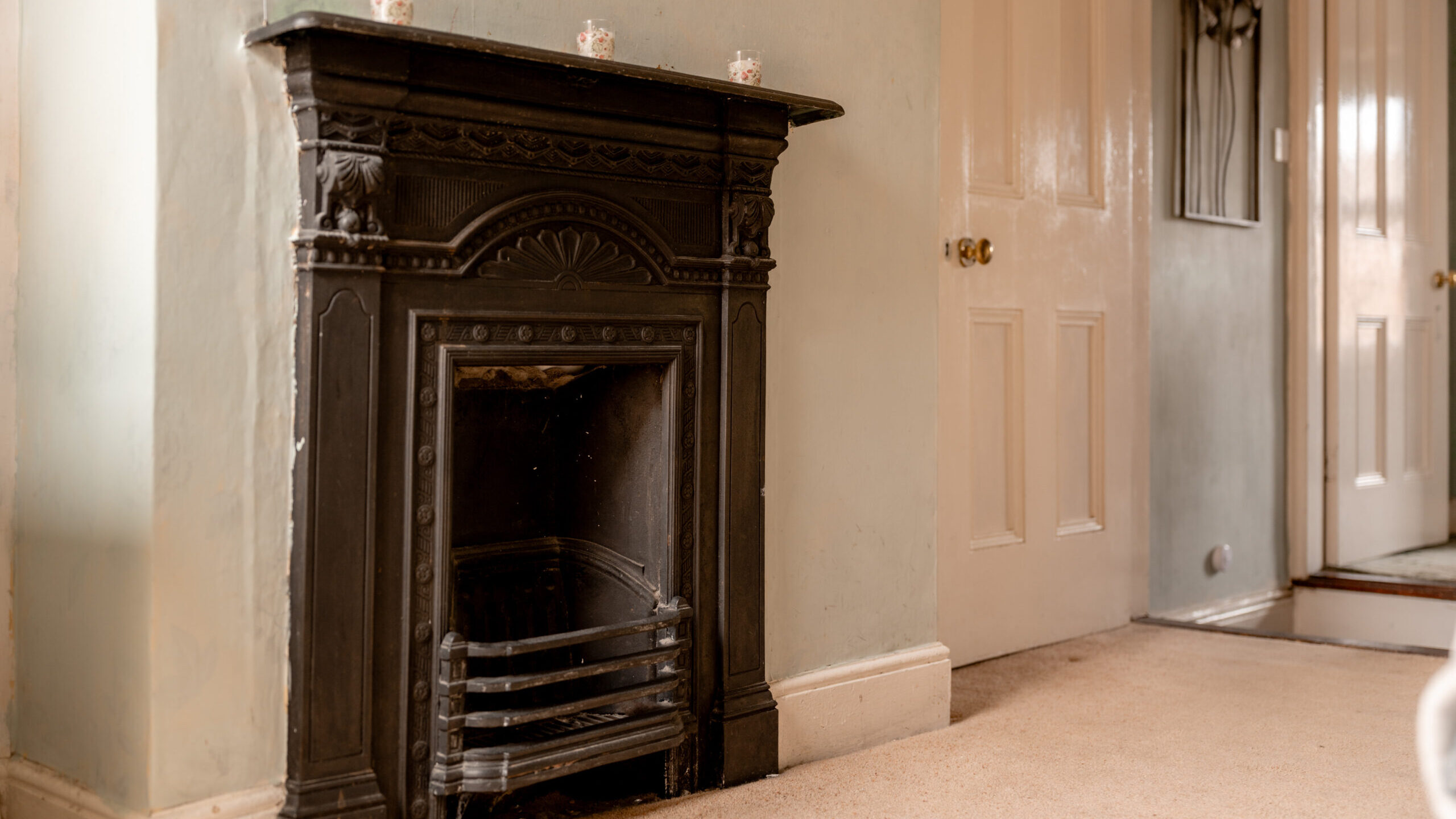 An ornate black cast-iron fireplace is set against a pale green wall. Three small decorative items sit on the mantel. Beige carpet covers the floor, and closed cream-colored doors are visible in the background.