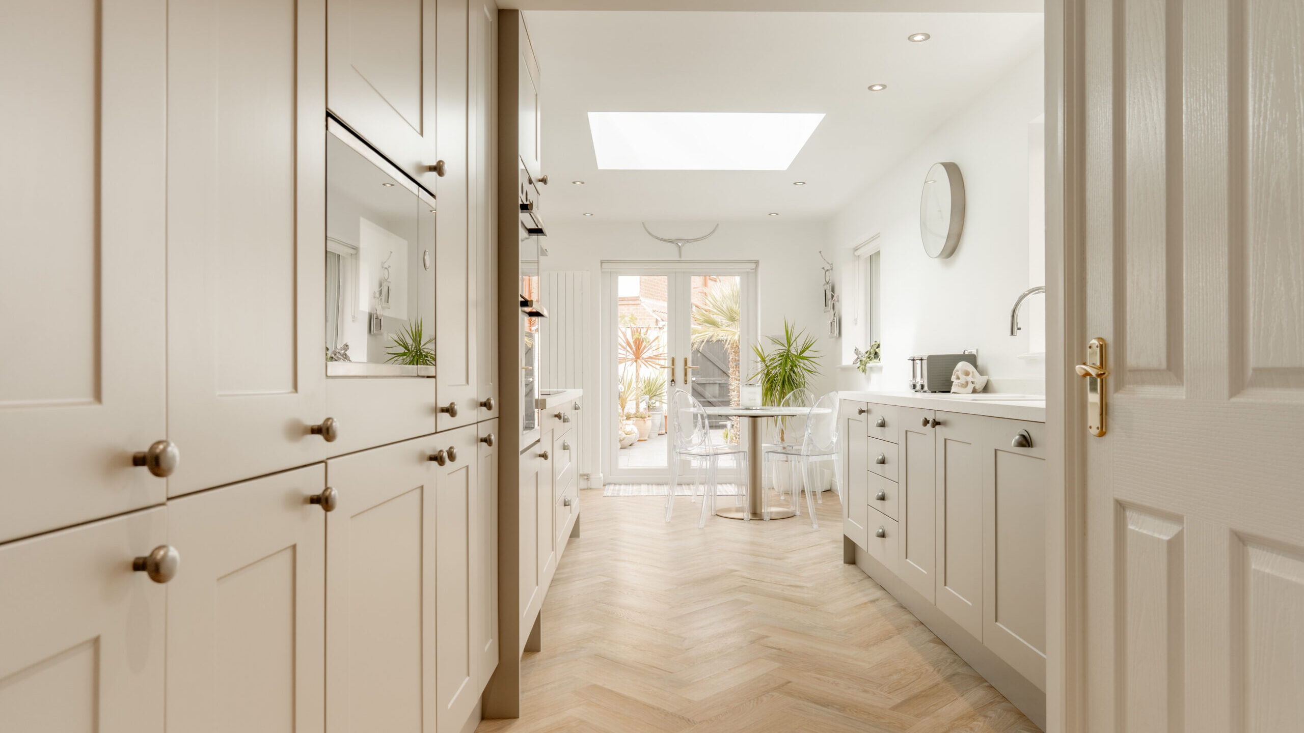 Bright modern kitchen with light beige cabinets, wood herringbone flooring, white countertops, and a skylight. A round glass dining table with chairs sits near large glass doors leading to a sunny garden.