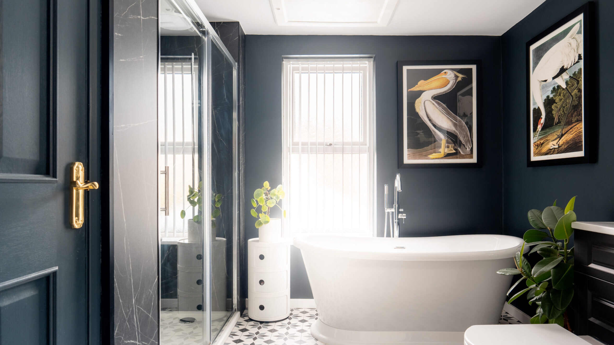 A modern bathroom with dark walls, a white freestanding bathtub, black and white patterned floor tiles, large shower, plants, and framed bird artwork on the walls. Vertical blinds cover the window.