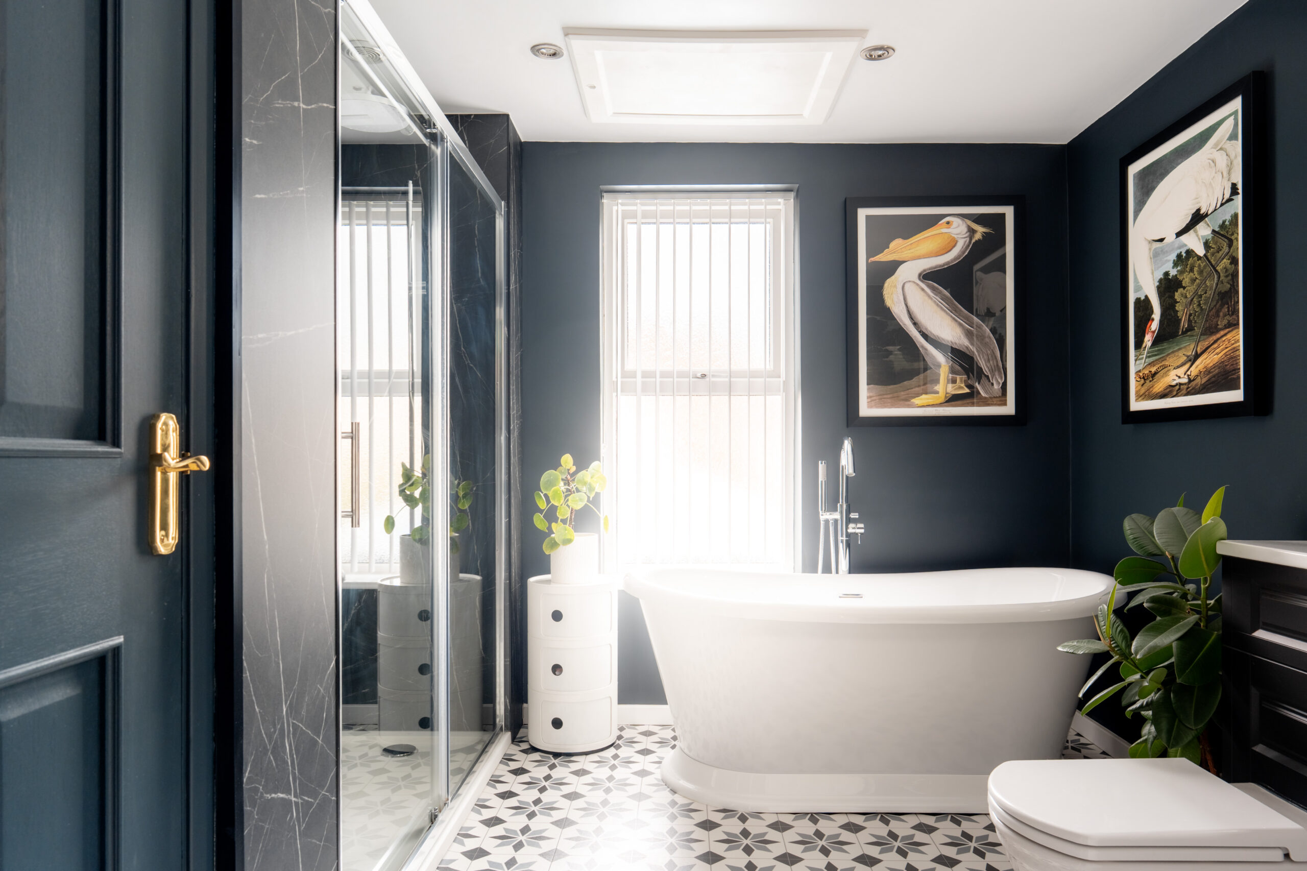 A modern bathroom with dark walls, a white freestanding bathtub, black and white patterned floor tiles, large shower, plants, and framed bird artwork on the walls. Vertical blinds cover the window.