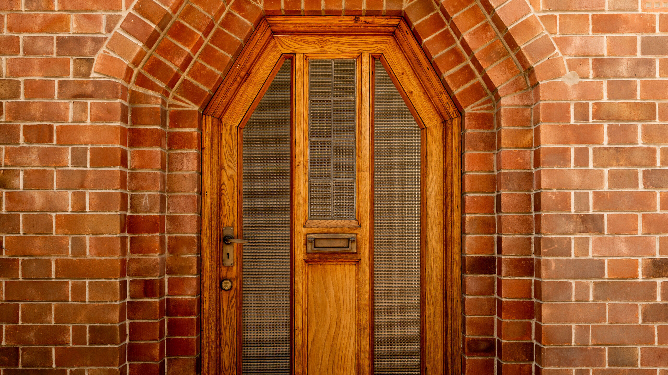 A wooden door with a mail slot, textured glass panels, and a black handle is set in a geometric brick archway with reddish-brown bricks. The bricks are arranged in a detailed, symmetrical pattern around the door.