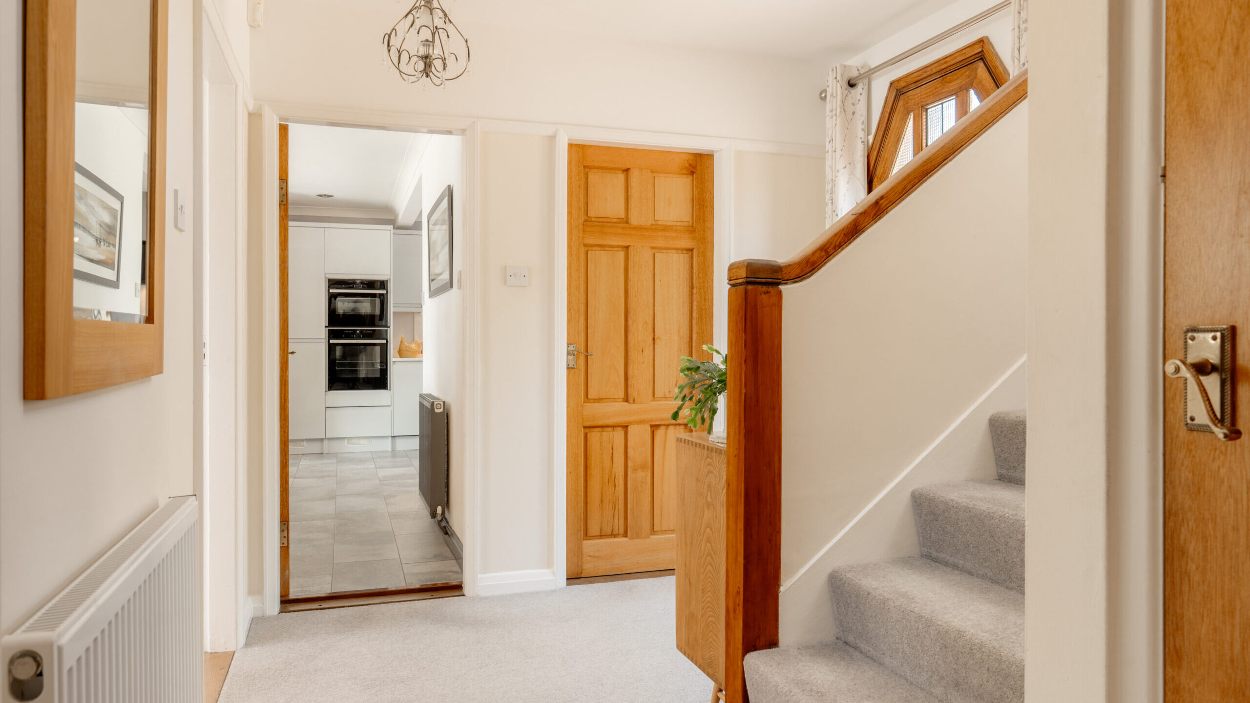 Bright hallway with light carpeting, wooden doors, a staircase with a wooden banister, and an ornate light fixture. The hallway leads to a modern kitchen with grey flooring and built-in appliances.