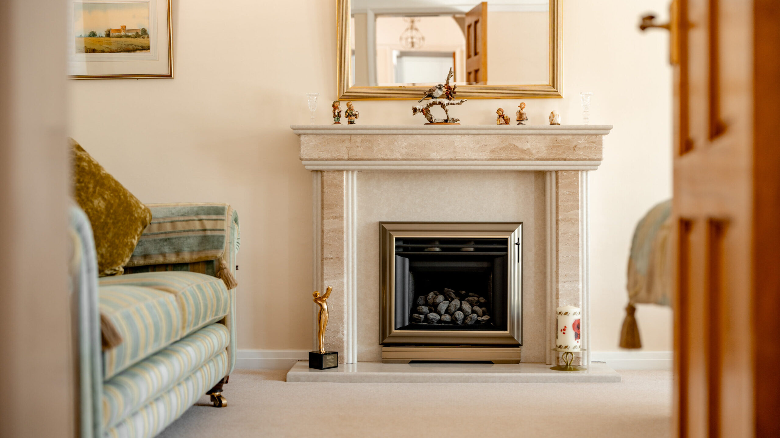 A cozy living room with a striped sofa, a decorative fireplace, a large mirror above the mantel, and small ornaments displayed on the mantelpiece. The room has soft, neutral colors and carpeted flooring.