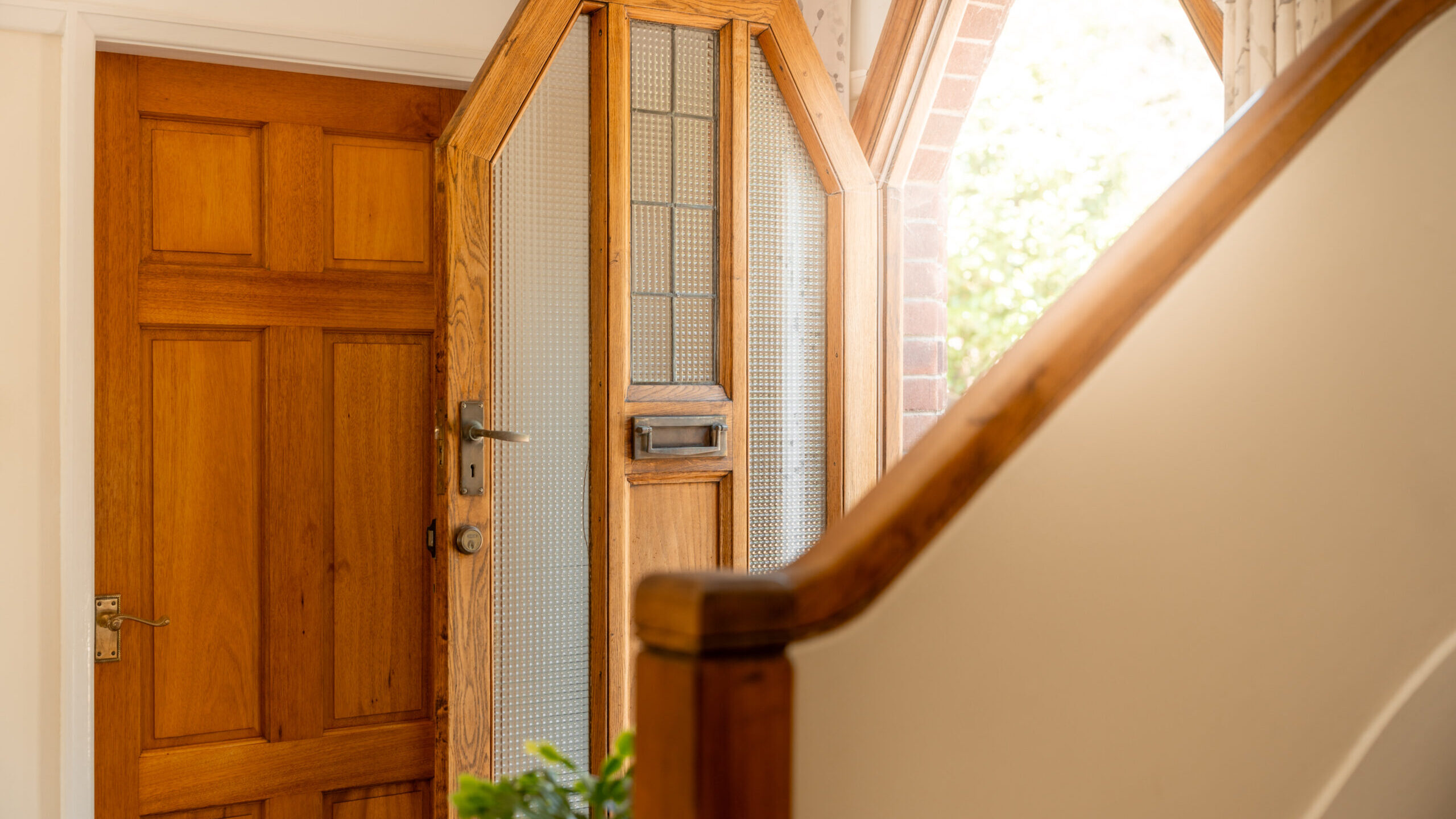 A bright entryway with a wooden front door, a partially open glass-paneled door, and a staircase with a wooden banister. Sunlight streams in through a window above the door.