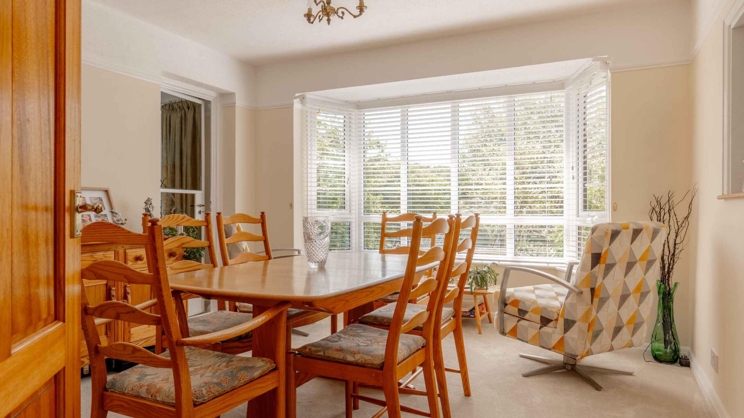 A bright dining room with a wooden table and six chairs, a patterned armchair by a large bay window with white blinds, neutral walls, light carpet, and a glass vase on the table.