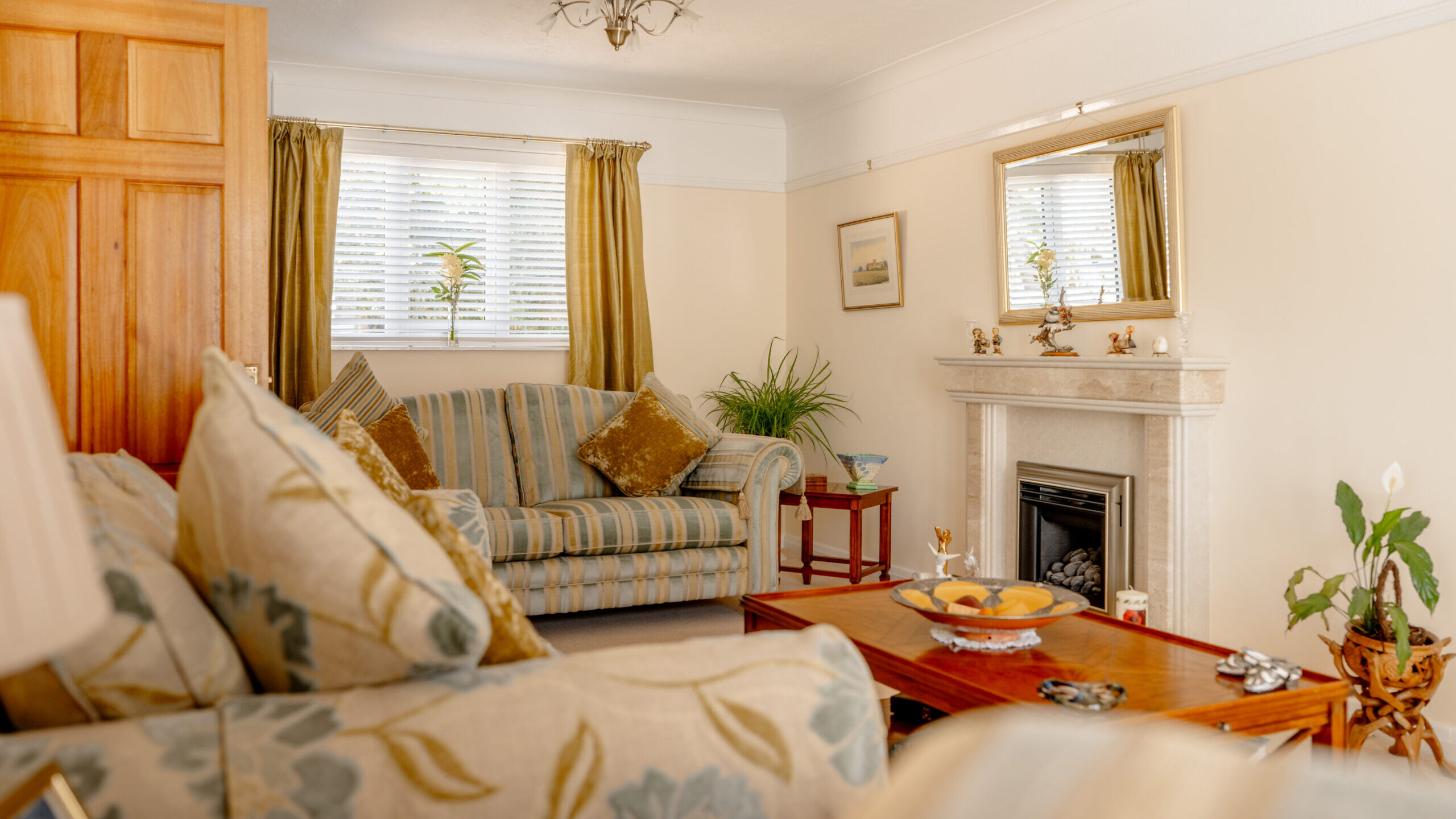 A cozy living room with striped and floral sofas, gold cushions, a wooden coffee table, a fireplace with a mirror above, potted plants, and golden curtains framing a window with blinds.