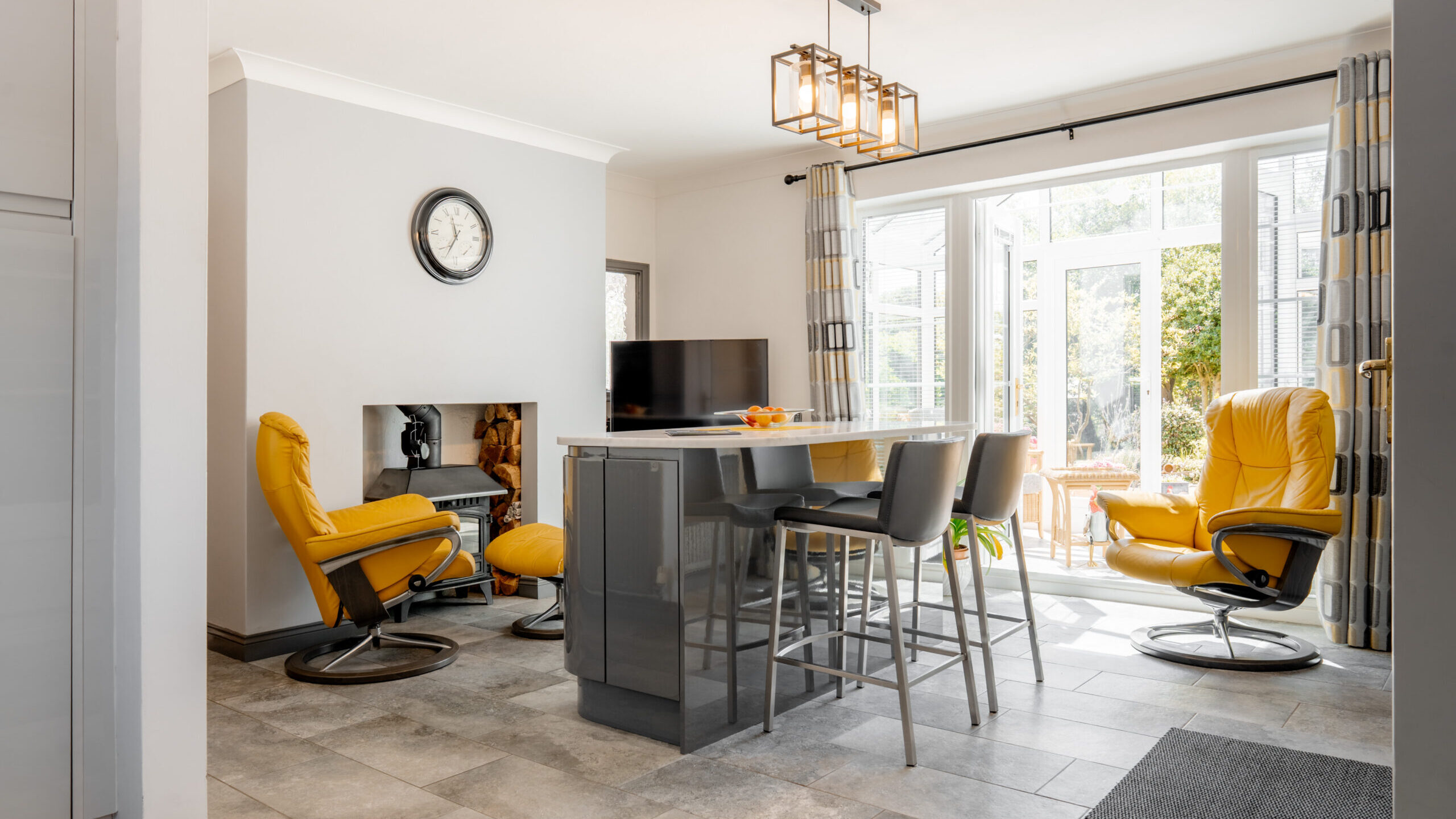 Modern kitchen and living space with gray tile floor, a central island with four gray chairs, two yellow armchairs, a wall clock, TV, and large windows letting in natural light with garden view.