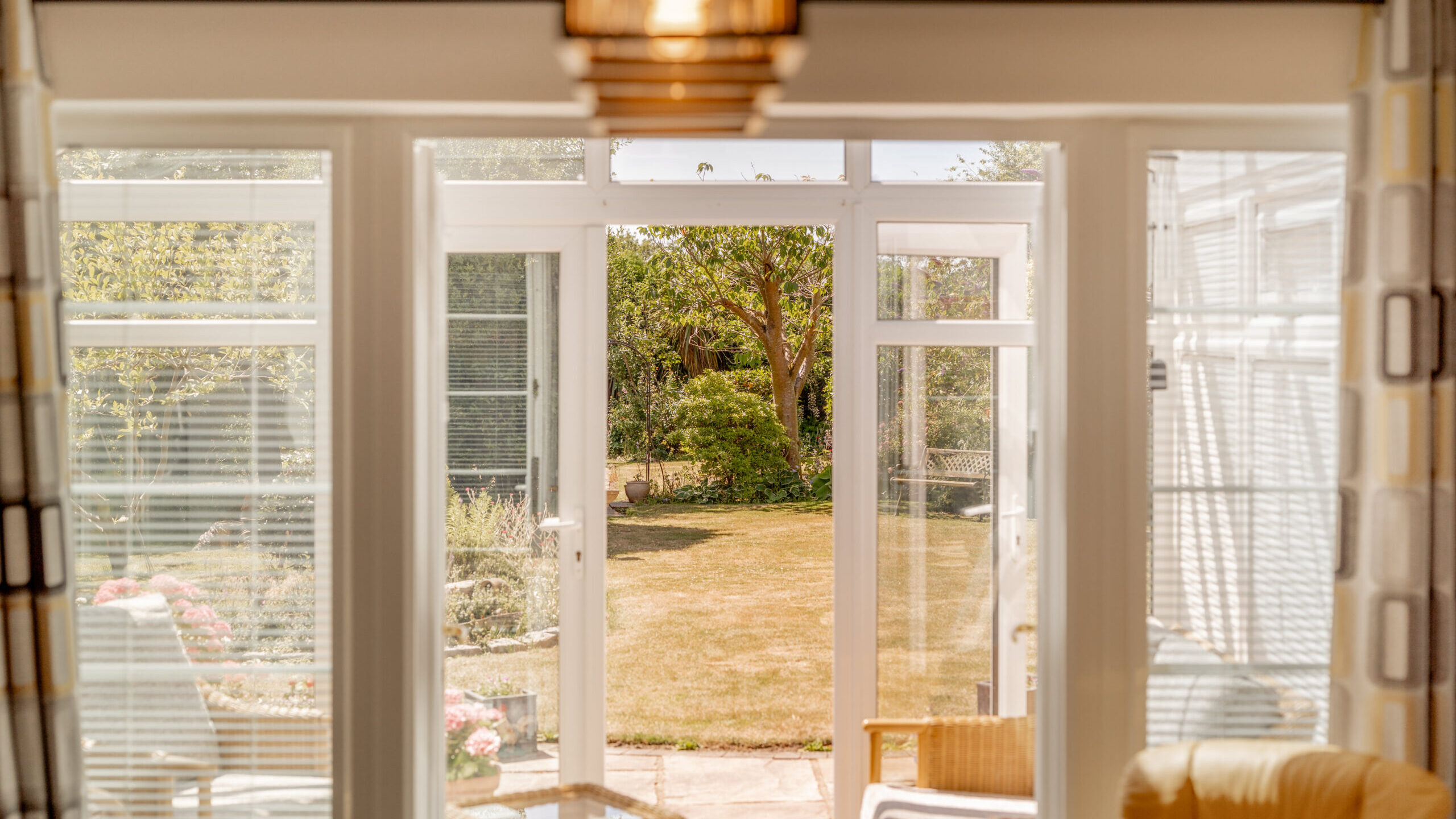 Sunlit conservatory with glass doors open to a garden, showing green grass, trees, and outdoor seating. Indoors, there are wicker chairs and a small table. Light streams in, creating a bright, inviting atmosphere.
