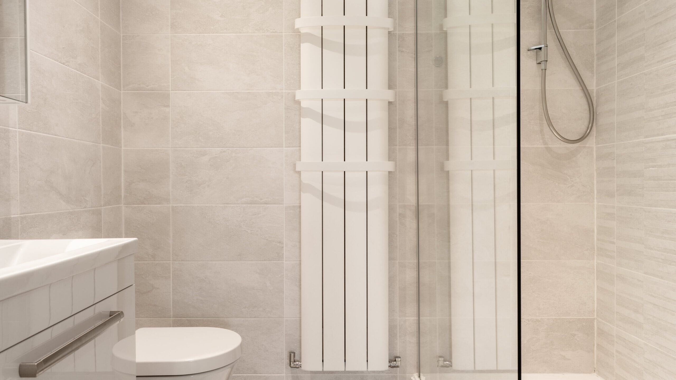 Modern bathroom with light gray tiles, a white wall-mounted toilet, a vertical white radiator, a glass shower enclosure, and a handheld showerhead. The space is clean, minimal, and well-lit.