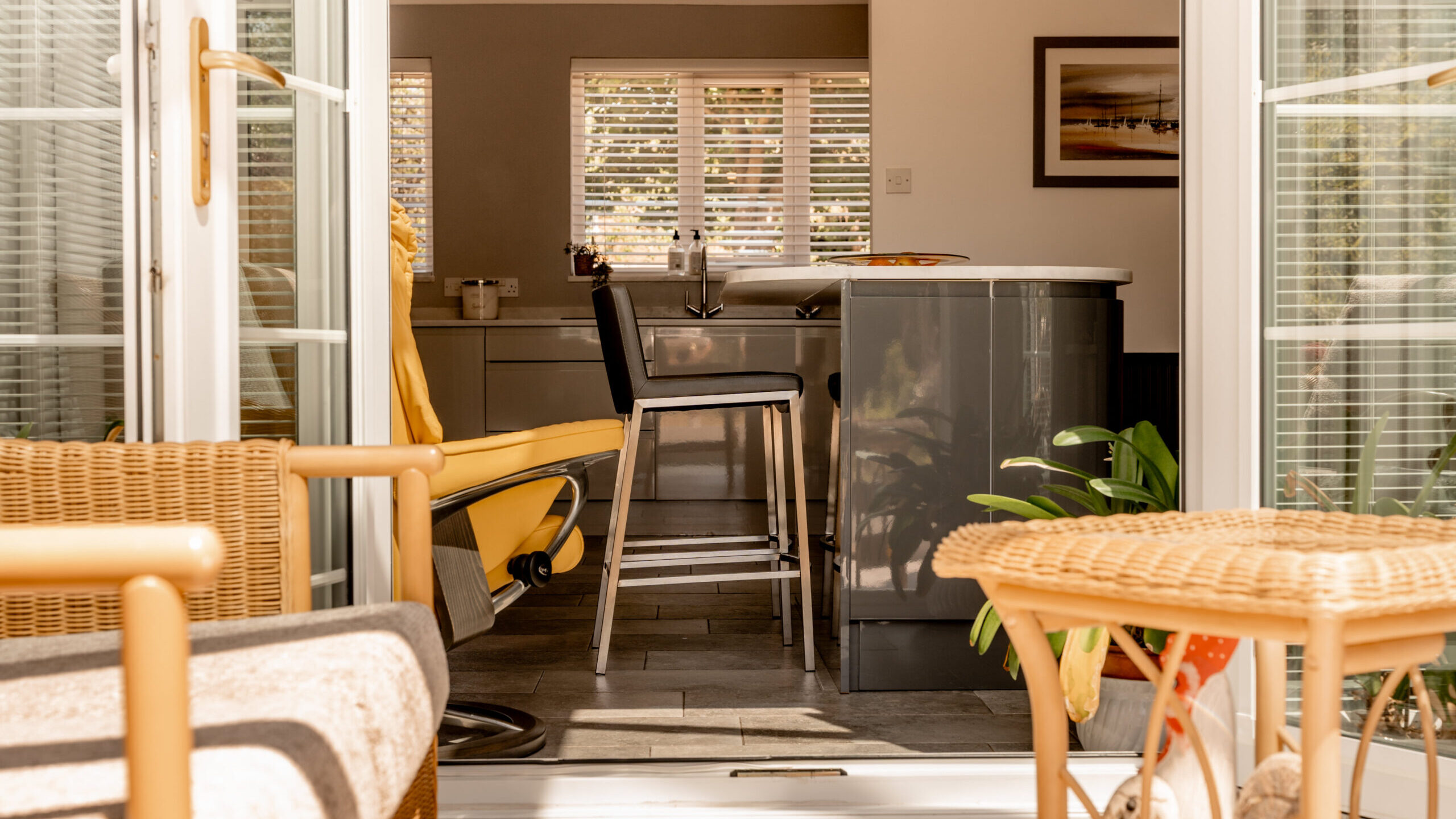 Sunlit modern kitchen seen through open double doors, featuring a central island with black chairs, a yellow armchair, wicker furniture, potted plants, and large windows with white blinds letting in natural light.