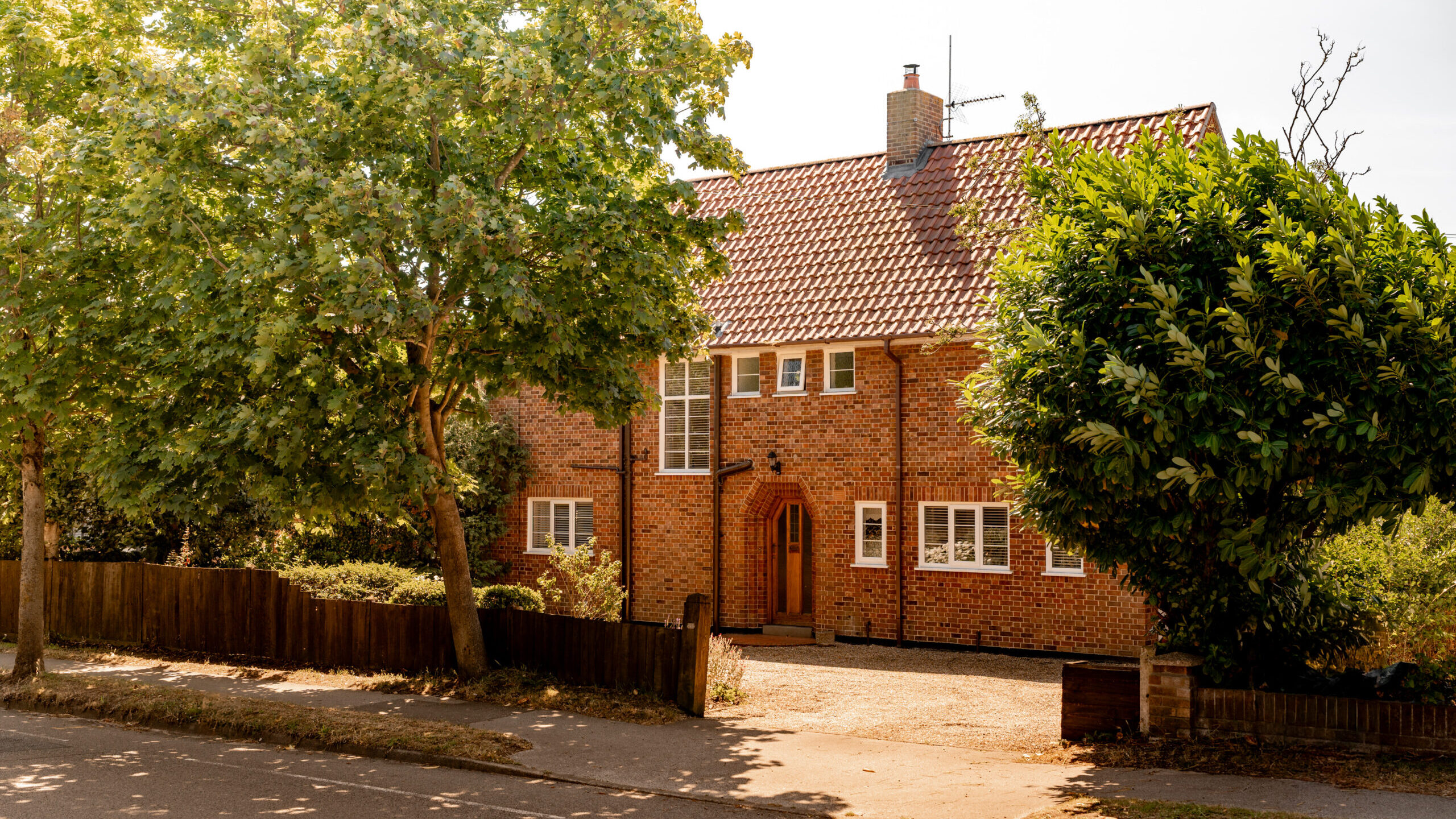 A two-story brick house with a red tiled roof, arched wooden front door, and large windows, surrounded by trees and a wooden fence, on a sunny day.