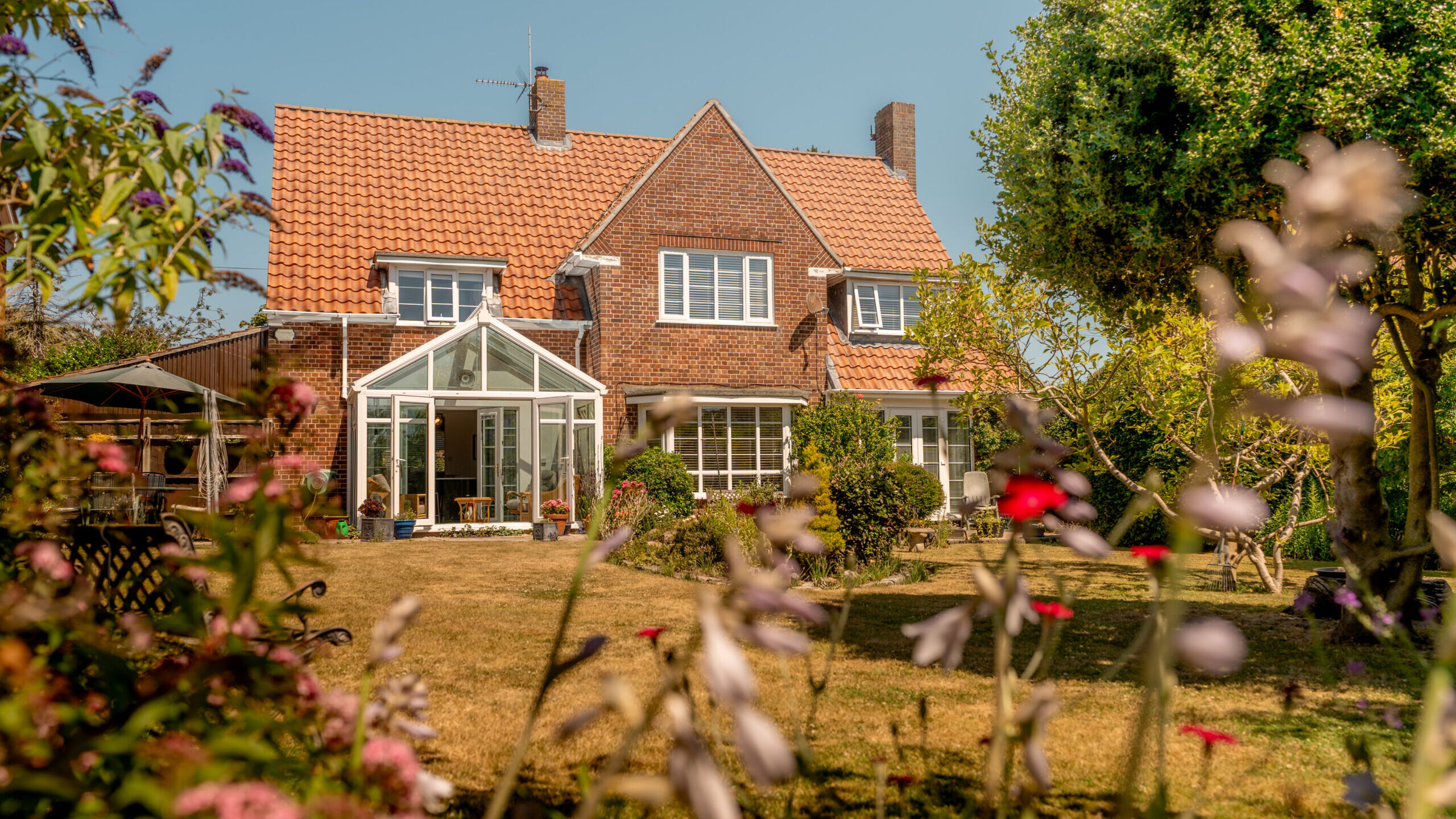 A red-brick house with a tiled roof and a glass conservatory sits in a sunny garden, surrounded by flowers, green grass, and trees under a clear blue sky.