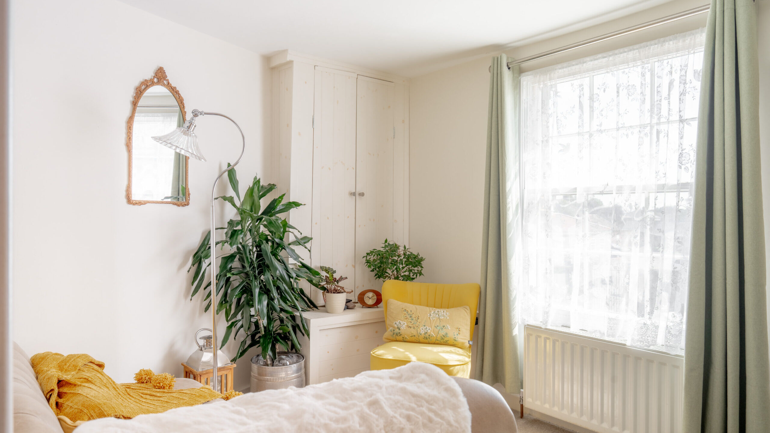 Bright living room with a beige sofa, yellow armchair, green plants, a wall mirror, and sheer curtains over a large window with green drapes. Light streams in, creating a cozy and inviting atmosphere.