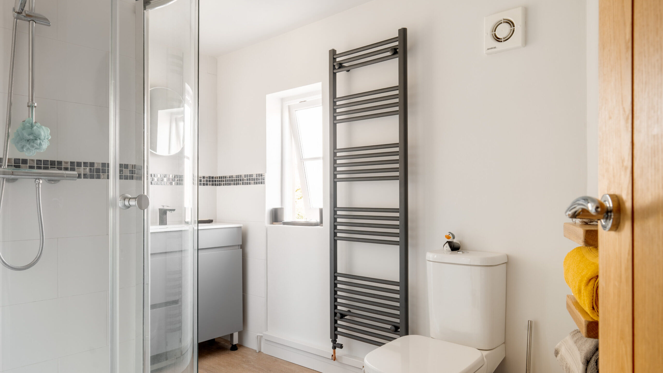 Modern bathroom with a glass shower, gray towel radiator, toilet, small sink with cabinet, wooden shelves holding towels, small frosted window, and white walls with light wood flooring.