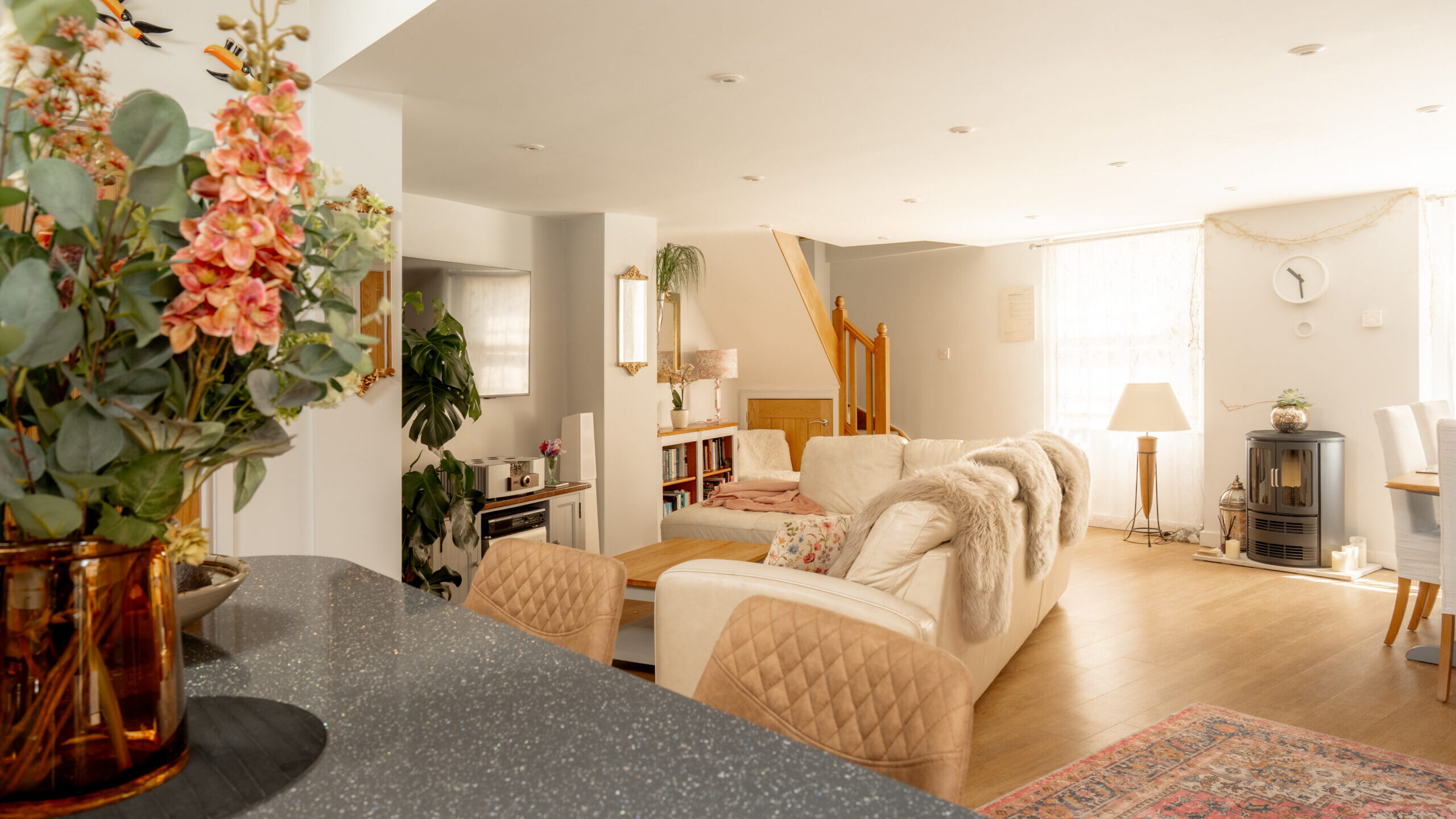 Bright, cozy living room with a white sofa, armchairs, and wooden floors. Large windows let in natural light. A vase with flowers sits on a counter in the foreground. Stairs and shelves are visible in the background.